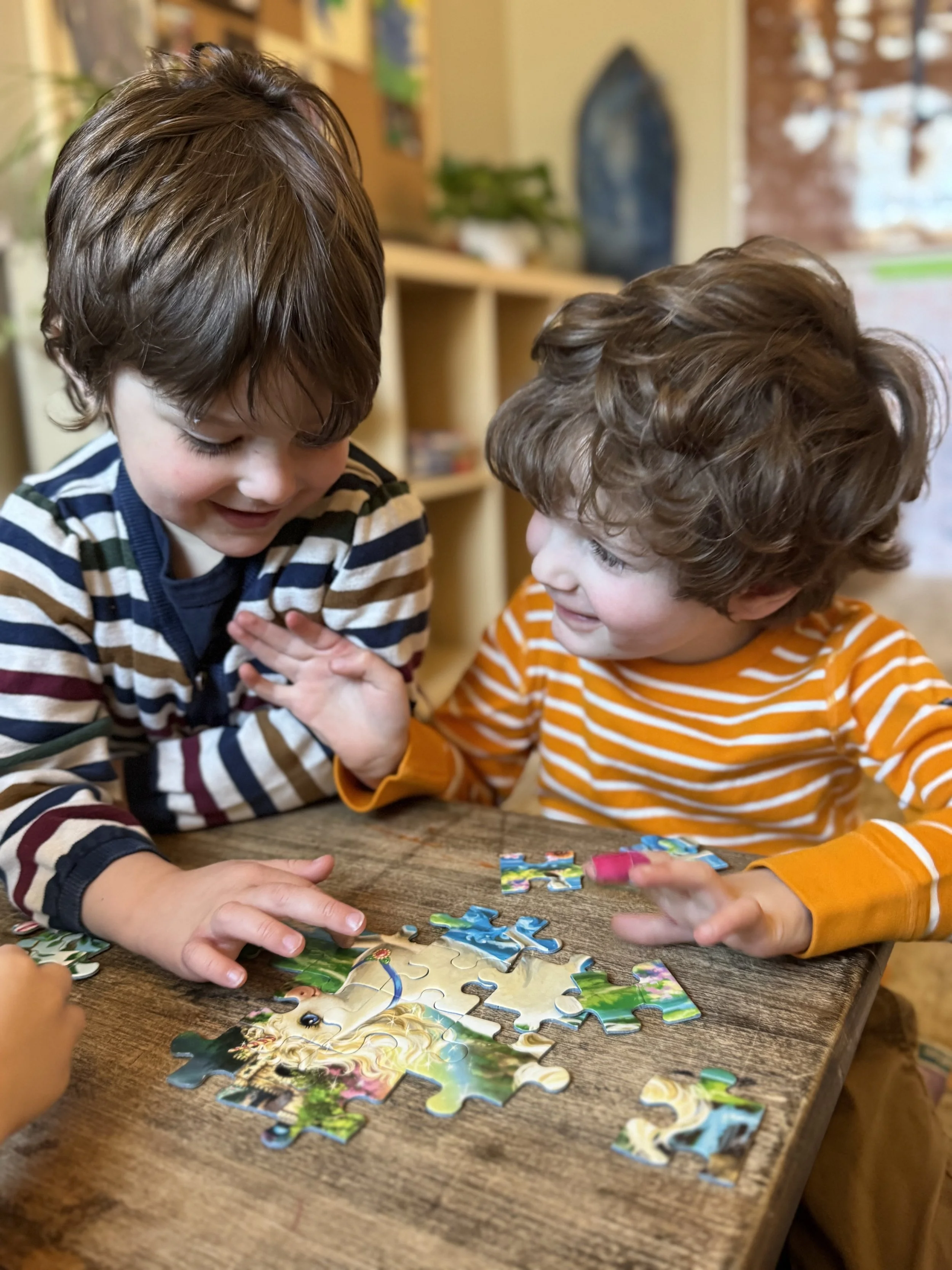 Two young children with light skin and curly brown hair smiling and playing with a puzzle on a wooden table.