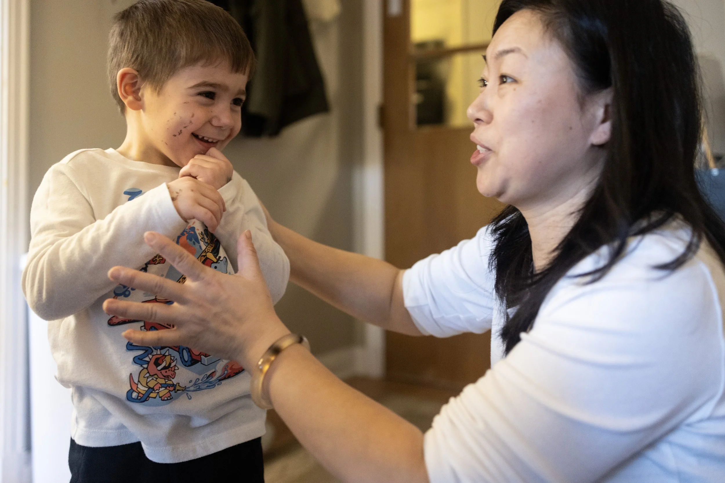 A woman and a young boy share a joyful moment inside a home. The boy is smiling and holding his chin with one hand, while the woman is reaching out to him with both hands, engaged in a cheerful interaction.