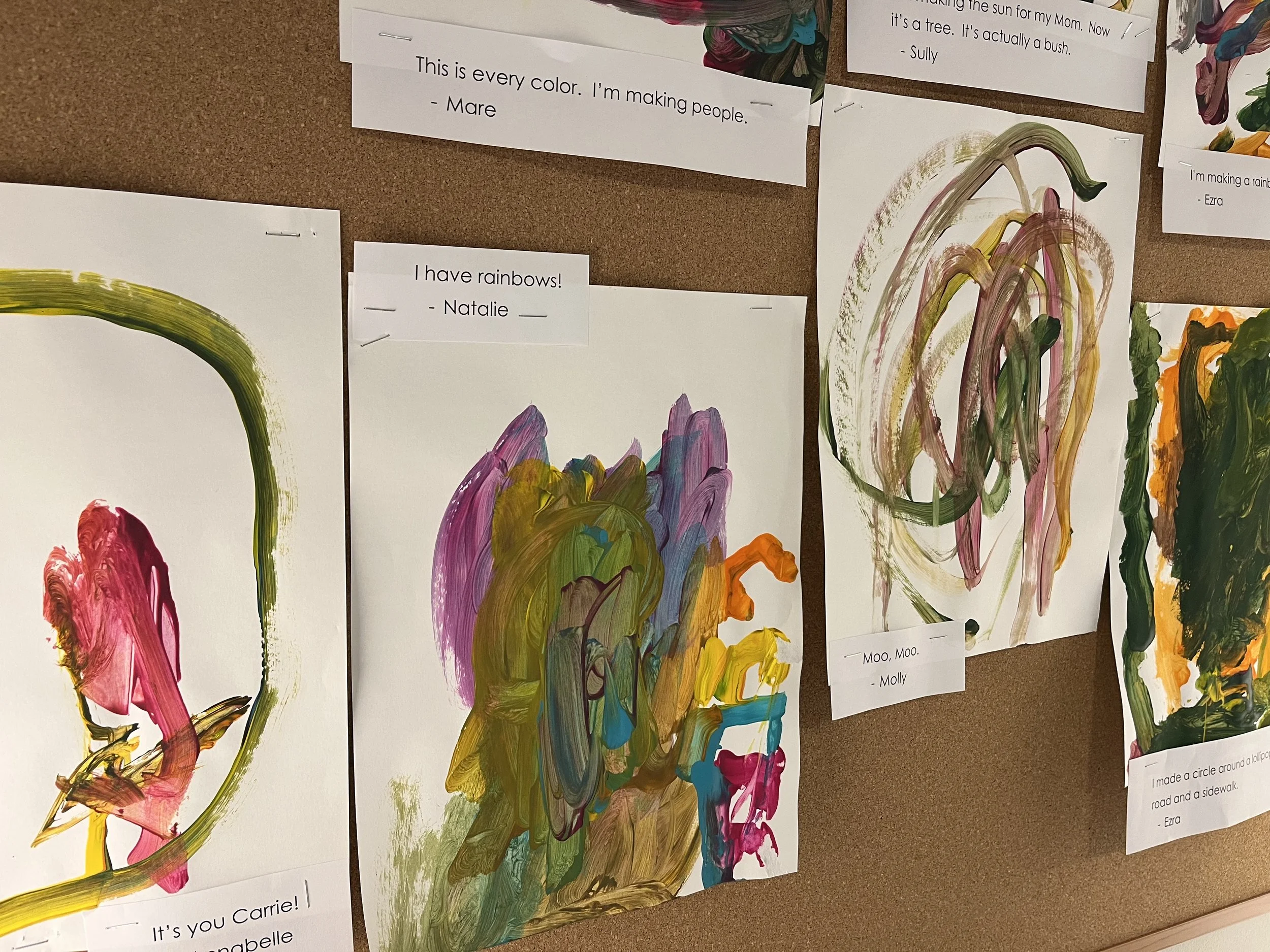 Collection of children's abstract paintings on paper, with colorful brushstrokes and notes with quotes and names attached to each artwork, displayed on a bulletin board.