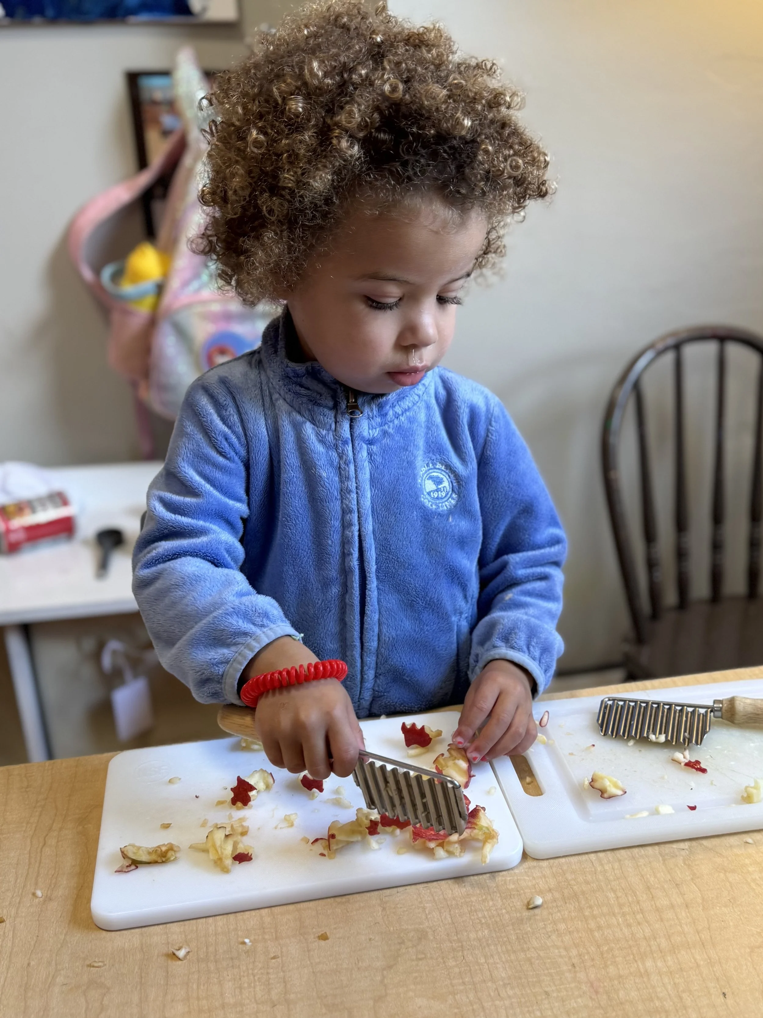 A young child with curly hair wearing a blue zip-up jacket, using a grater to shred a piece of food on a white cutting board. There are pieces of food on the table and a second empty cutting board nearby.