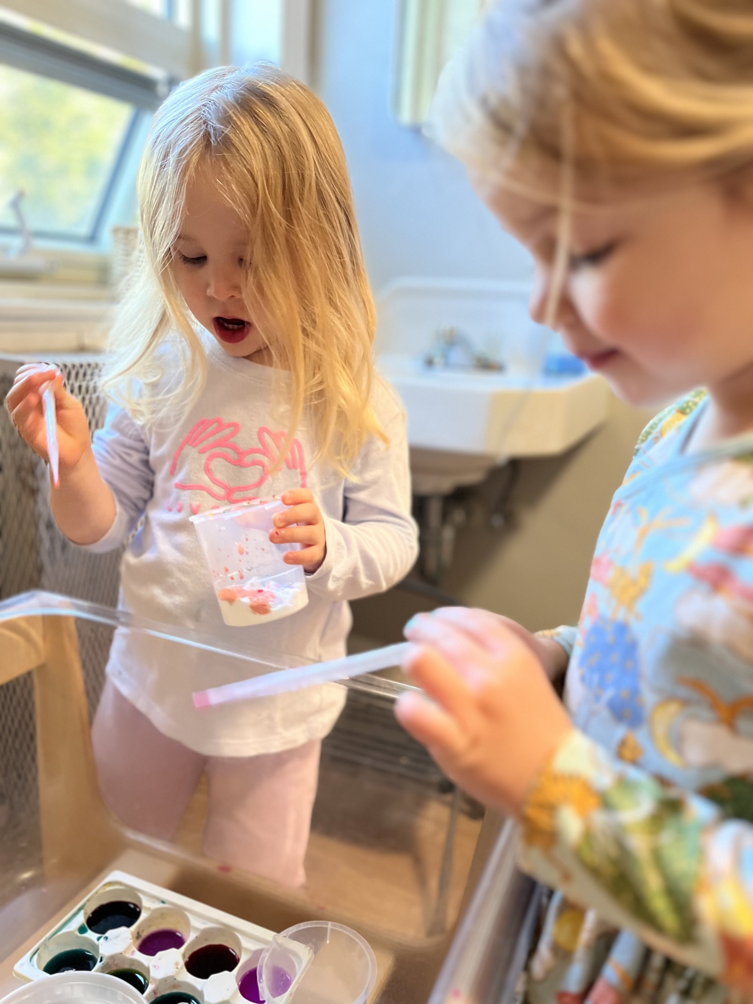 Two young girls painting with watercolors, one holding a cup and the other holding a brush, in a bright room.