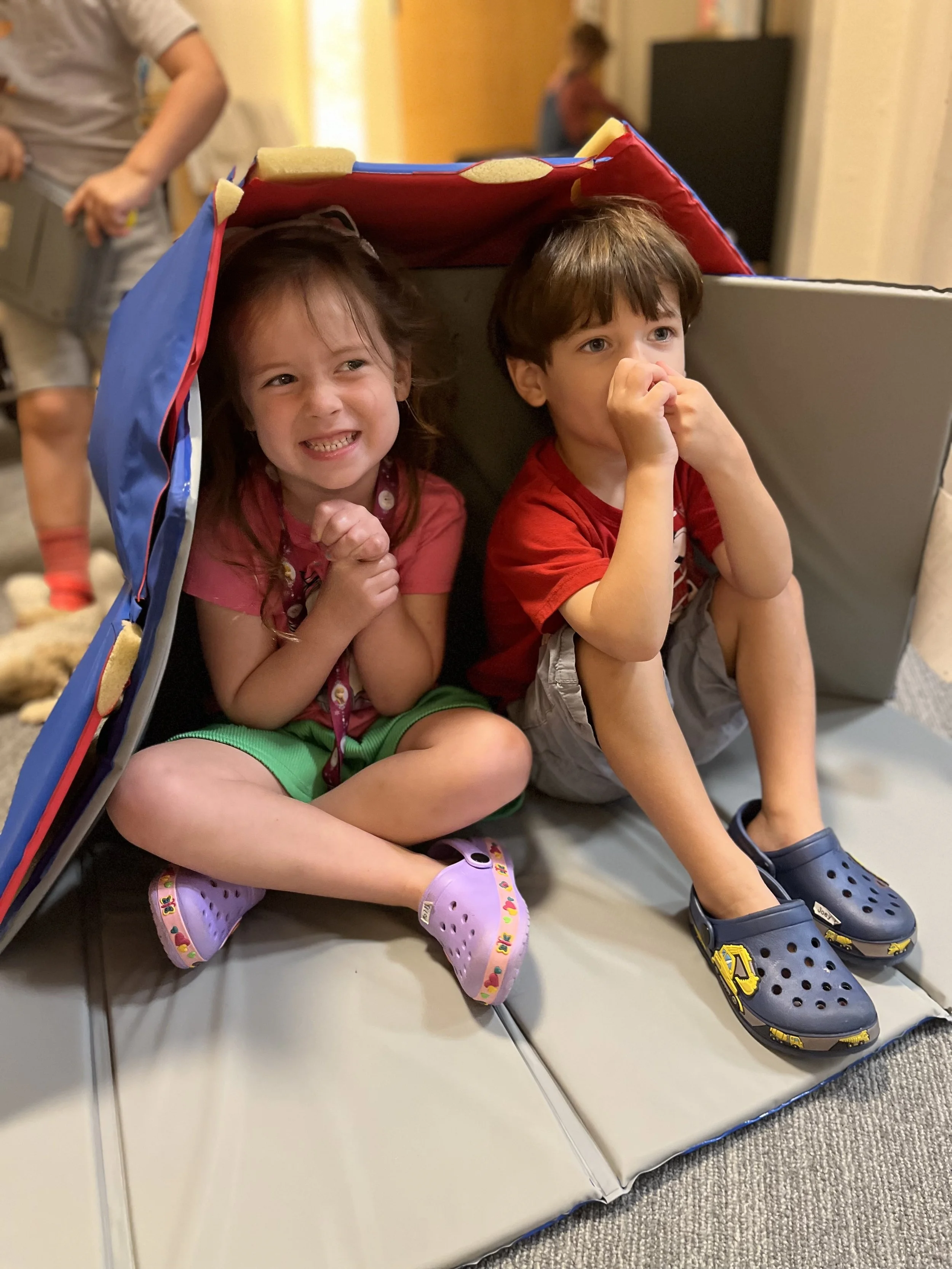 Two young children, a girl and a boy, sitting inside a large cardboard box with a roof made from a rolled-up blanket or mat. The girl is smiling and has her hands clasped in front of her, wearing a pink shirt, green shorts, and purple Crocs. The boy appears anxious or shy, holding his nose with both hands, wearing a red shirt, light-colored shorts, and blue Crocs with yellow accents. In the background, an adult is visible holding a bowl or container, and another child is sitting further away in the room.