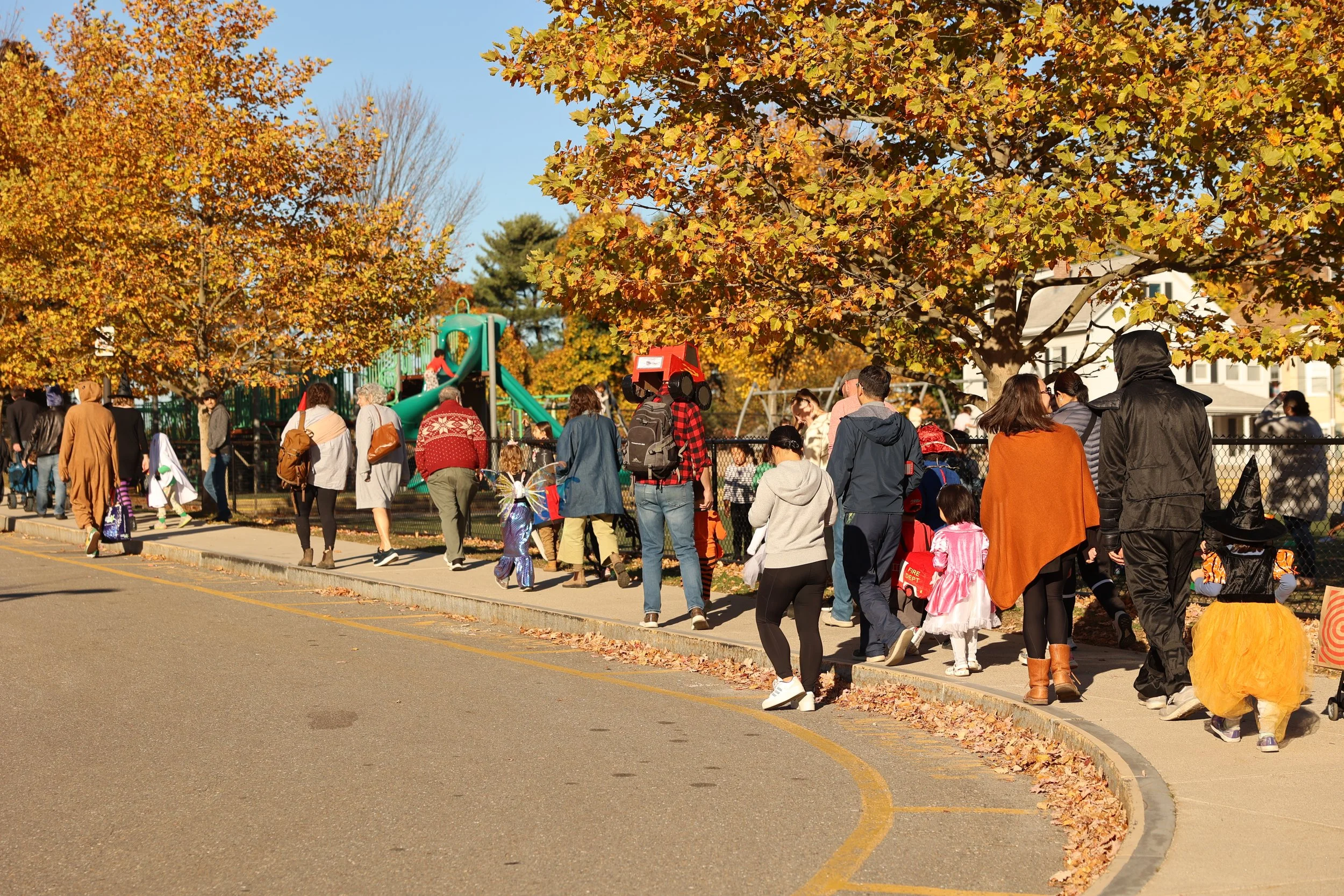 People dressed in costumes walking in a line near a playground with trees displaying autumn leaves.