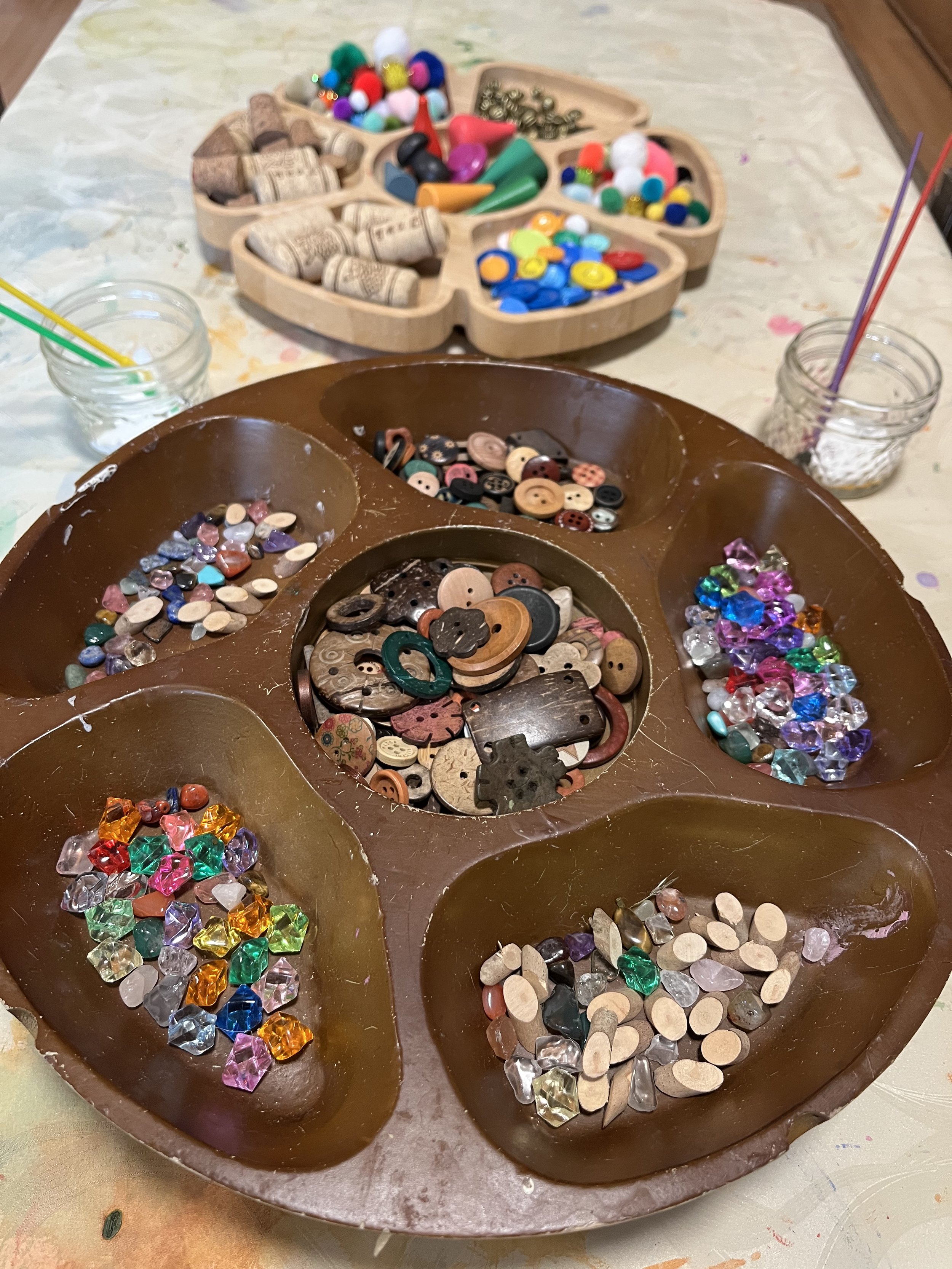 A paint palette filled with various buttons and craft items, with containers of colorful beads, pins, and pins in the background.