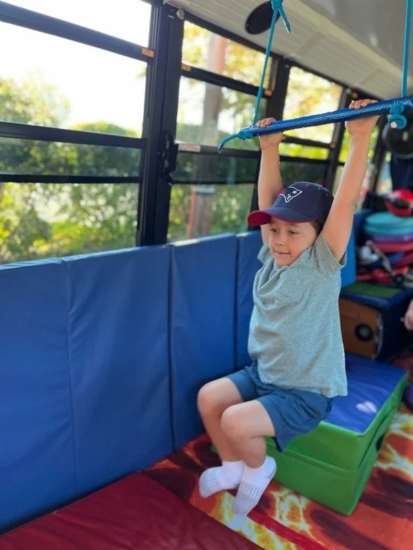 A young boy wearing a baseball cap, gray T-shirt, and shorts is hanging from a blue gymnastic bar inside a bus or play area. He is holding onto the bar with both hands, with his legs bent and feet off the ground, smiling and enjoying the activity.