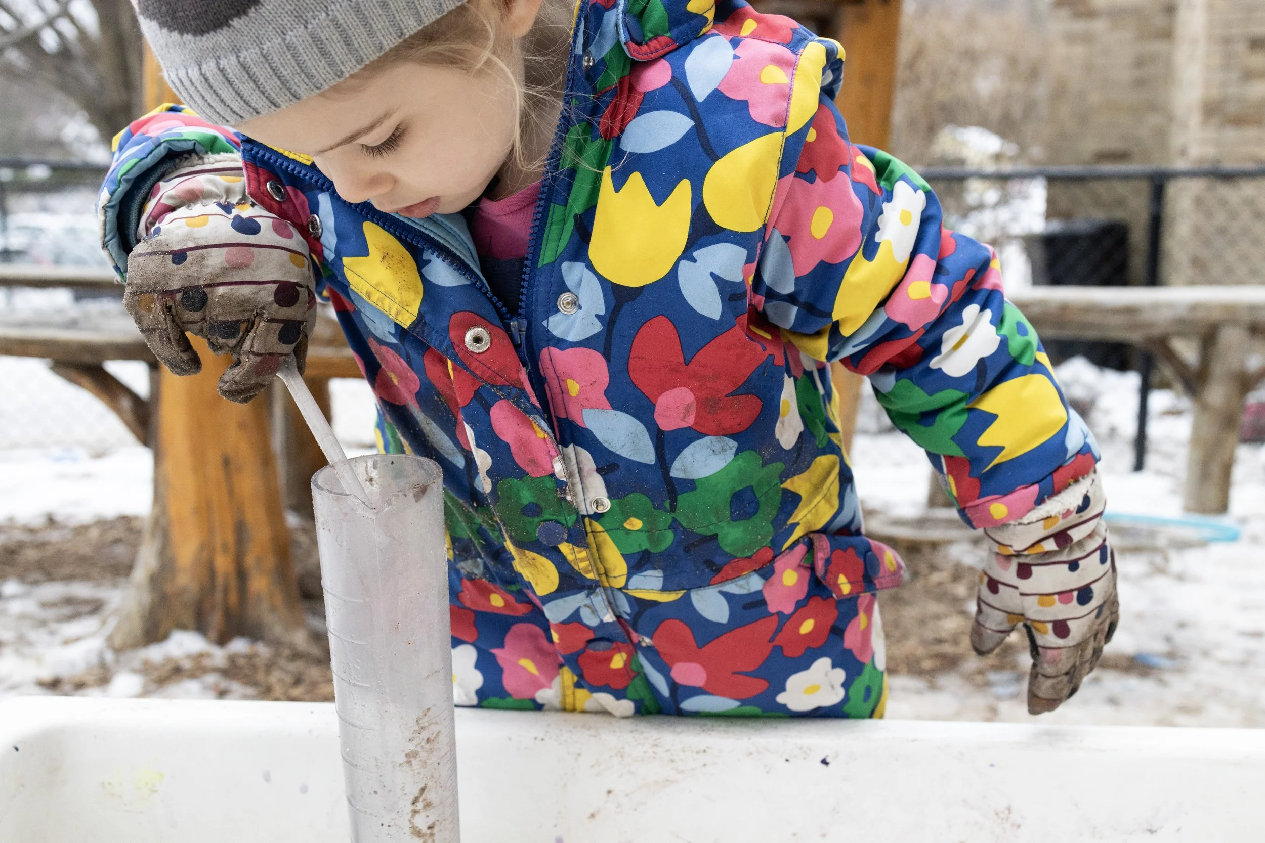 Child in a colorful floral winter coat and gloves playing outdoors in the snow, handling a clear plastic tube.