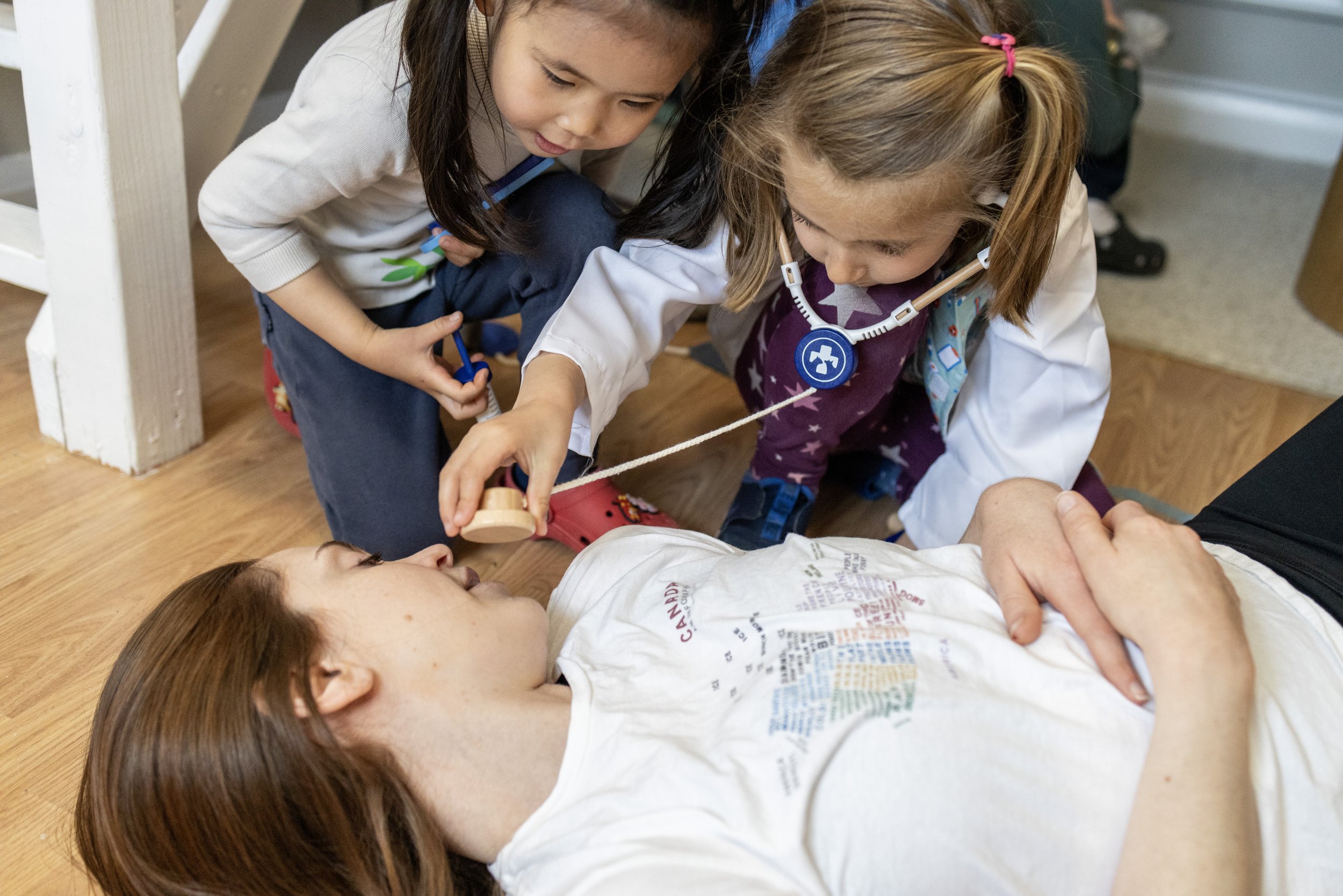 Children playing with a toy doctor kit while pretending to check on a woman lying on the floor, simulated as a patient.