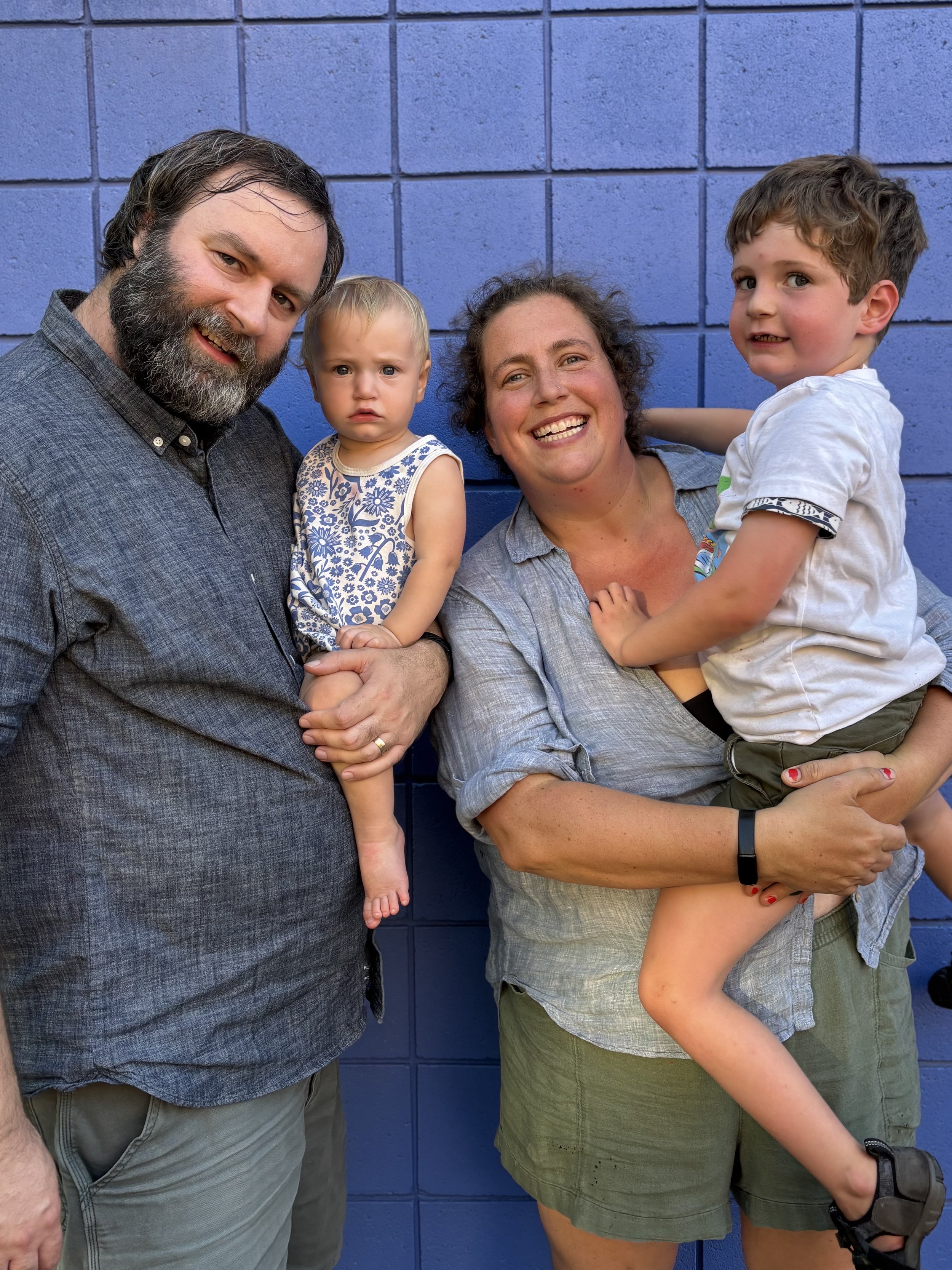 A family of four posing together against a blue wall. The father has a beard, the mother is smiling, and their young son is being held by the mother. Their young daughter stands next to the father, with a serious expression.