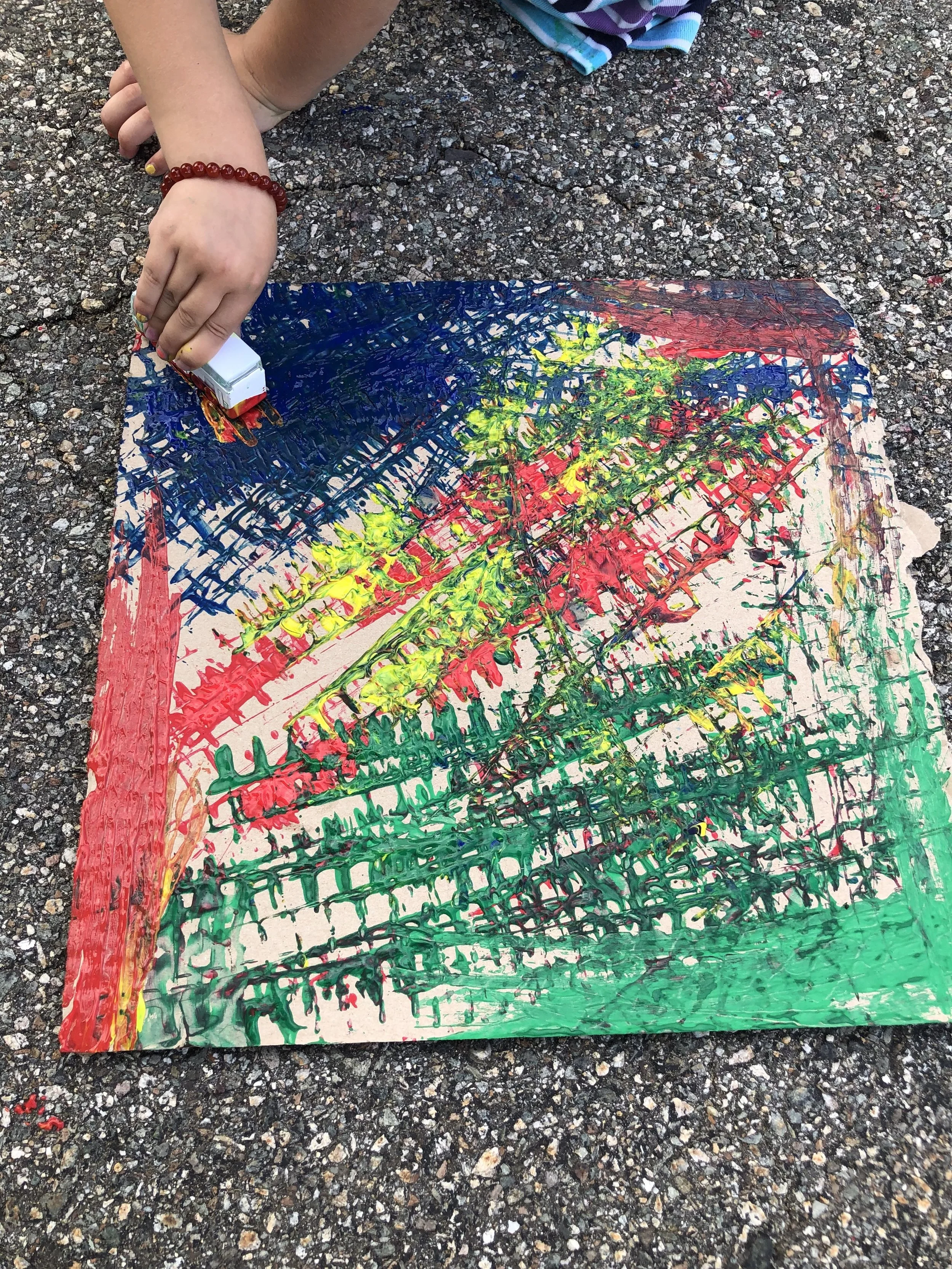 Child creating a colorful abstract painting on a piece of paper laid on asphalt, using various colors of paint and a small box of paint.