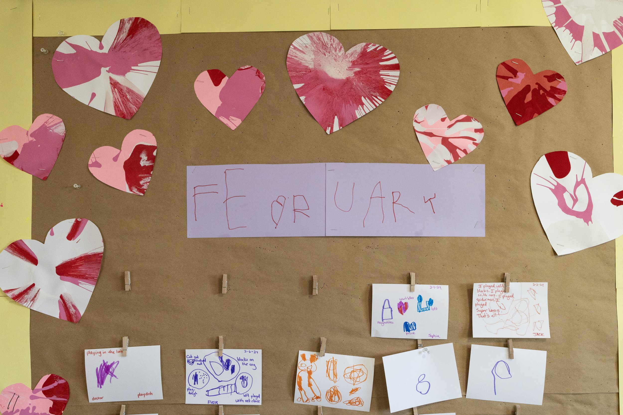 A bulletin board decorated with paper hearts made of red, pink, and white paint with abstract patterns. In the center, there is a sign with the message 'FEAR YOUR.' Below, small children's drawings are clipped to the board with clothespins, featuring colorful scribbles, animals, and figures.