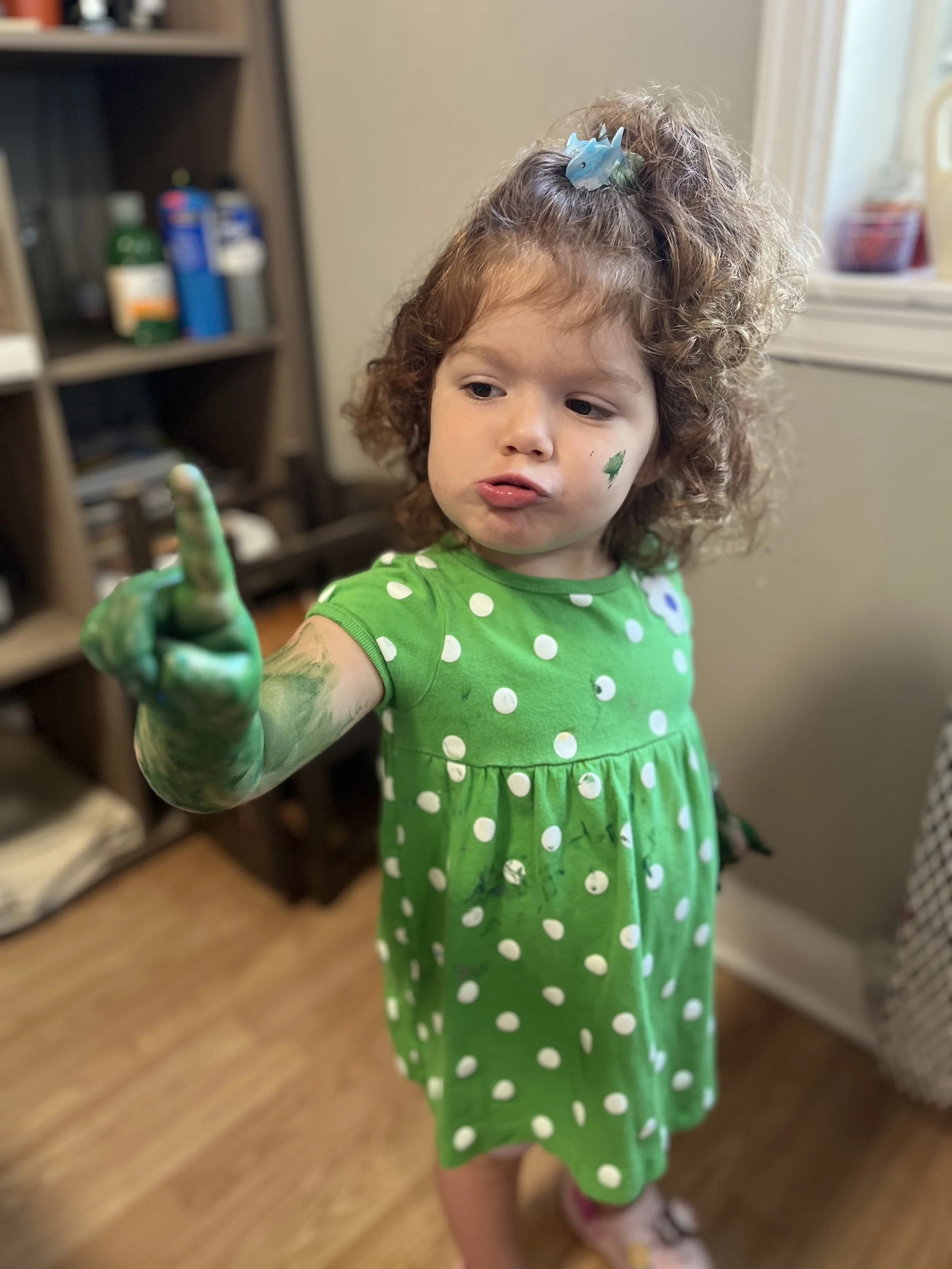 A young girl with curly hair wearing a green dress with white polka dots, covered in green paint on her arm and hand, pointing with her finger, with some paint on her face, standing indoors near a bookshelf and a window.