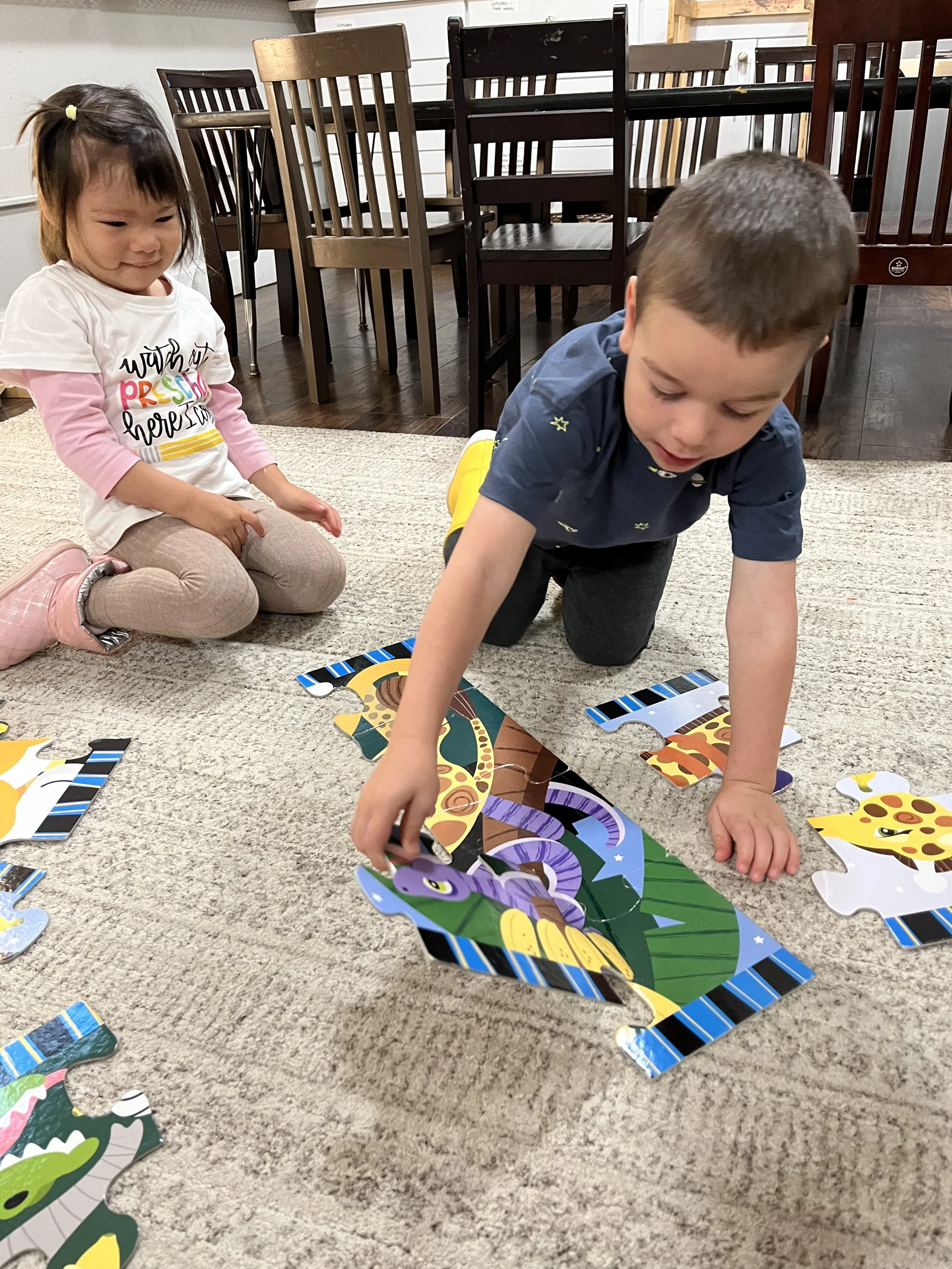 Two young children, a boy and a girl, sitting on a beige carpet in a room with wooden chairs, working on colorful animal-themed jigsaw puzzles.