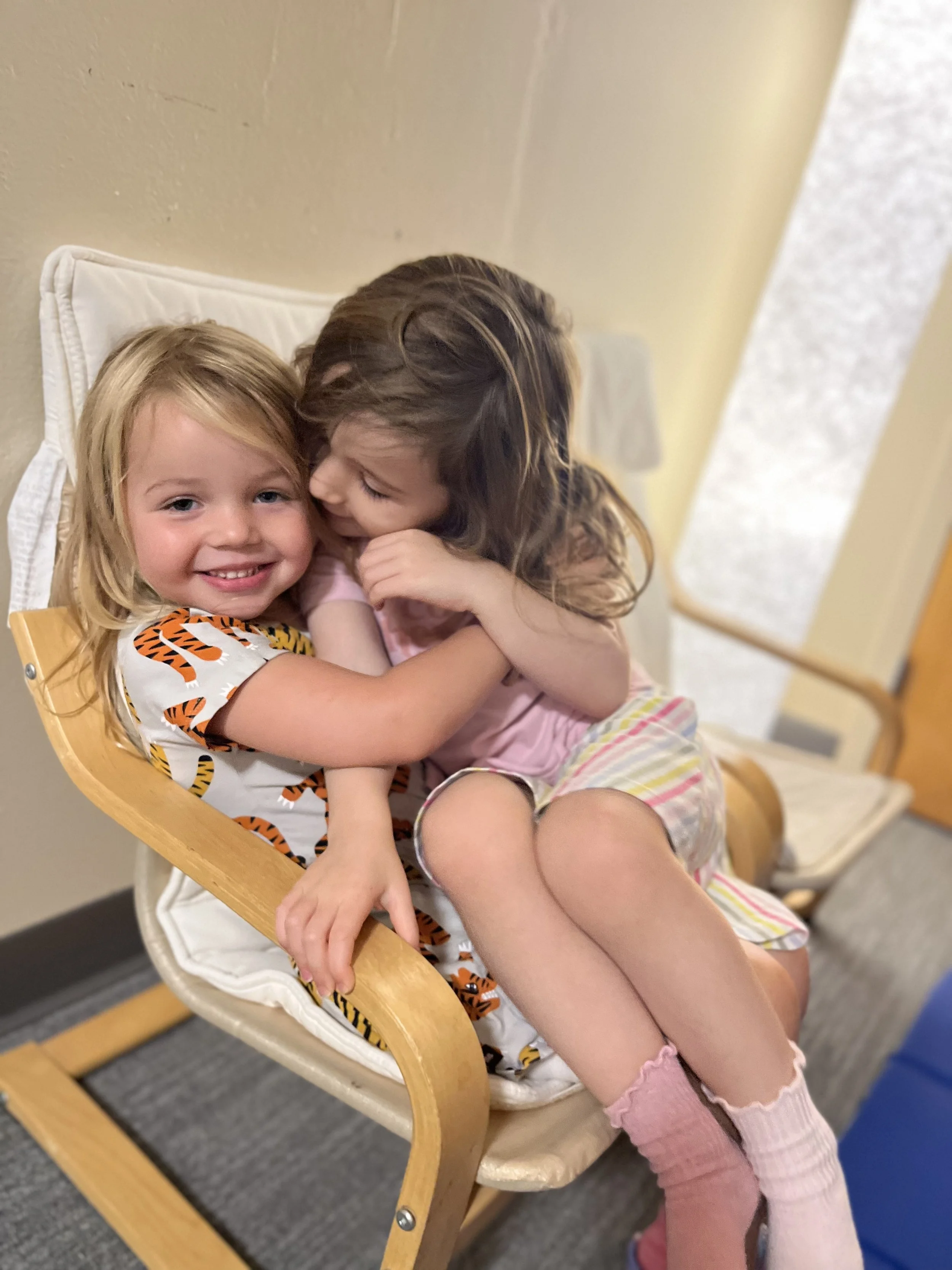 Two young girls hugging and smiling while sitting on a beige and wooden chair in an indoor setting.