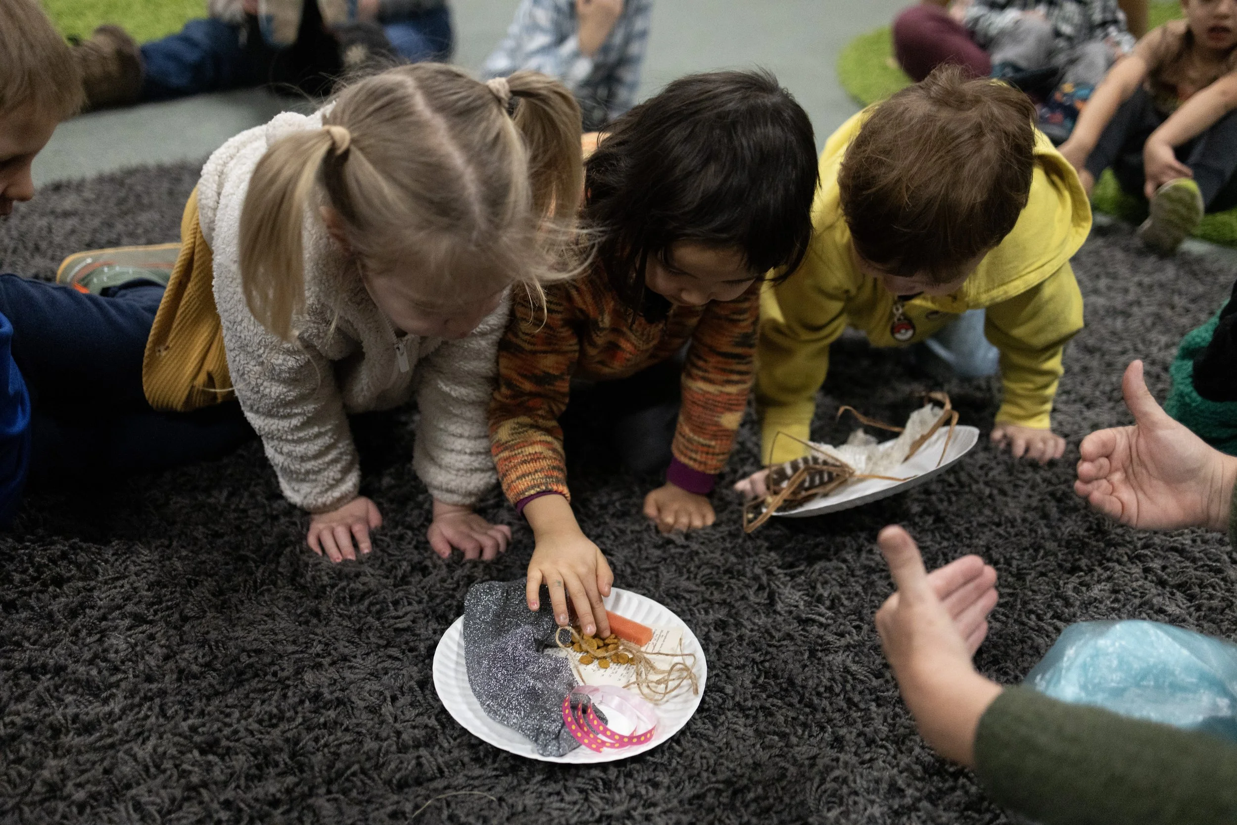 Children participating in a hands-on activity, examining objects on paper plates while on a carpeted floor, with other children sitting in the background.