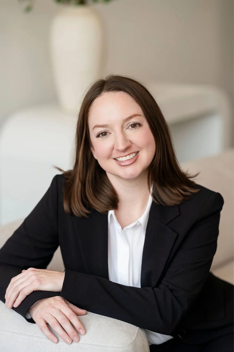 A smiling woman with shoulder-length brown hair, wearing a black blazer and white blouse, sitting at a table with her arm resting on the surface in a well-lit room.