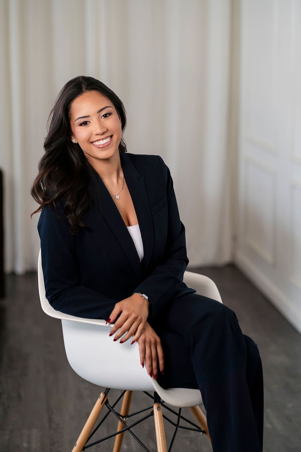 A woman dressed in a black blazer and white top sitting on a modern white chair with wooden legs in a room with white curtains and walls.