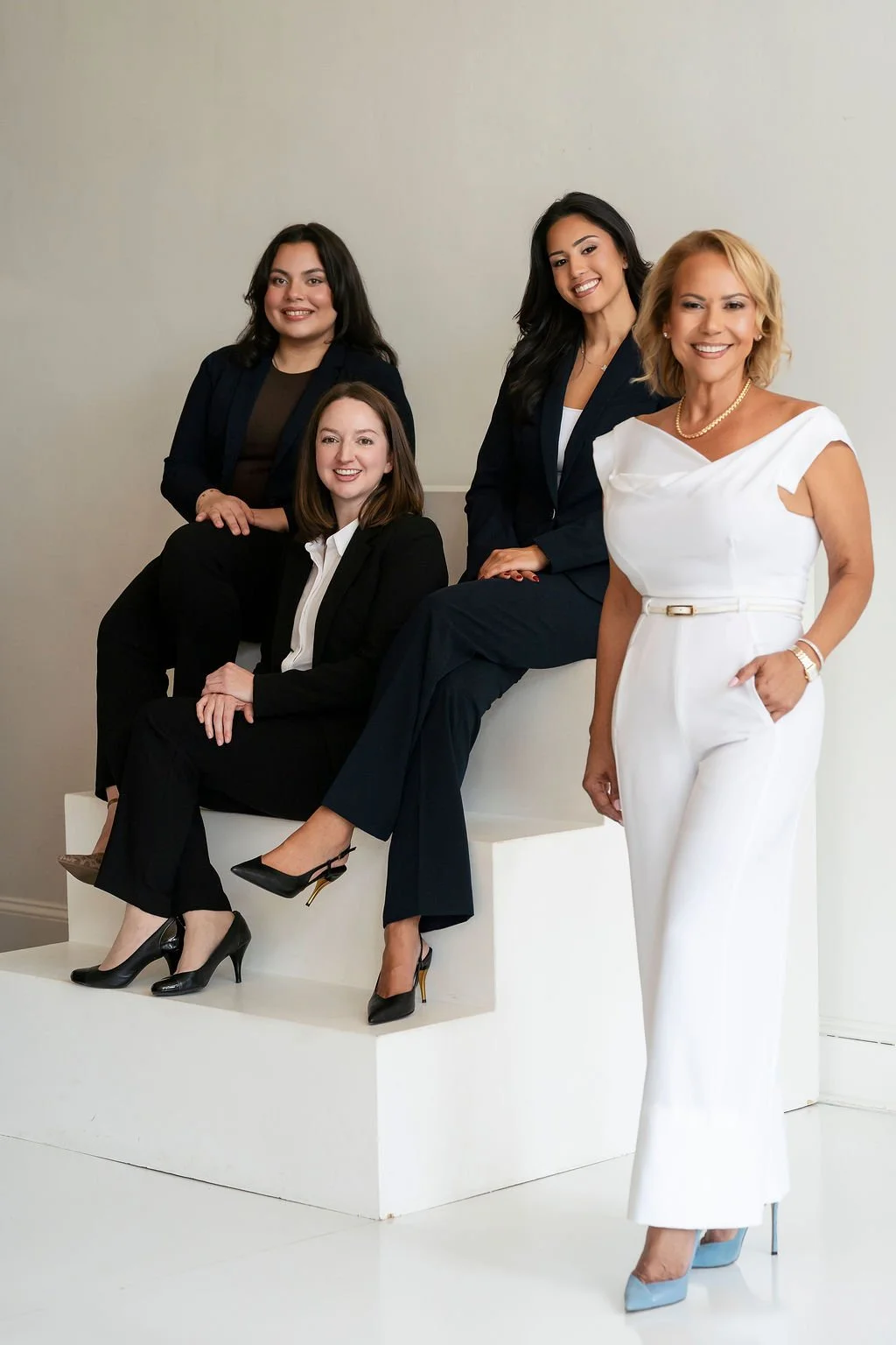 Group of five women dressed in professional attire posing together indoors against a plain background.