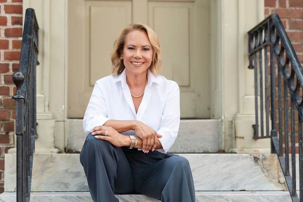 A smiling woman with blonde hair, wearing a white blouse, seated on steps outside a brick building with a black iron railing.