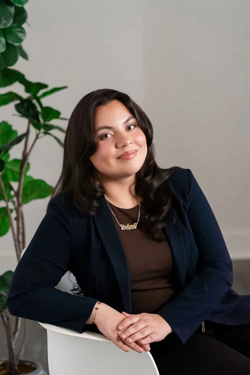 A woman with long dark hair, wearing a brown top, a dark blazer, and a necklace, sitting on a white chair with her hands folded, smiling at the camera, with a green plant in the background.