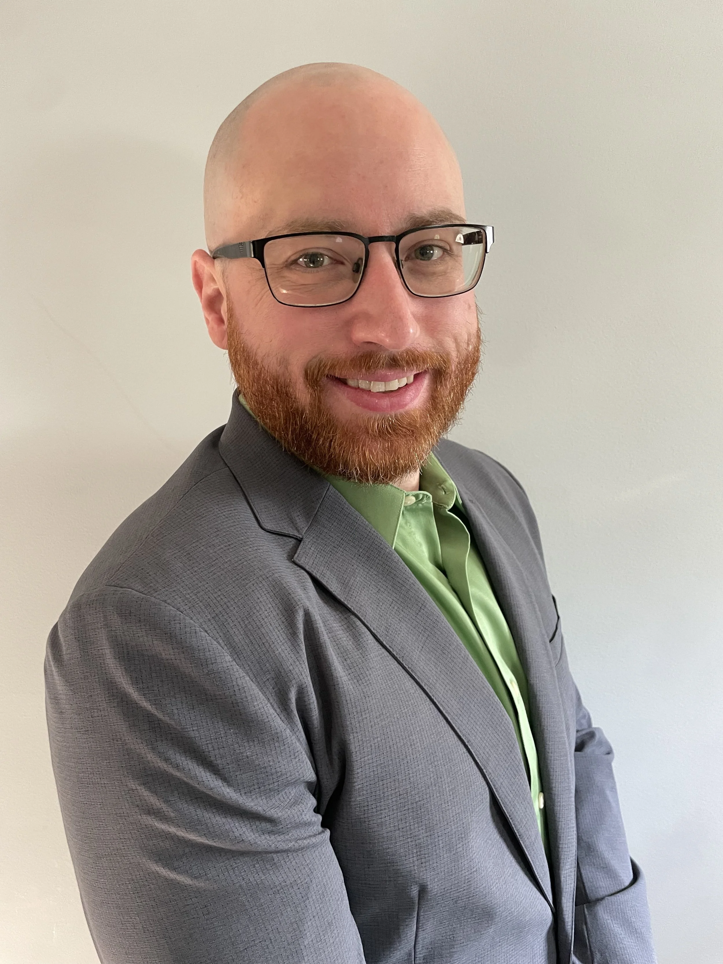 Headshot photo of Natan, wearing glasses and a grey blazer, smiling at the camera