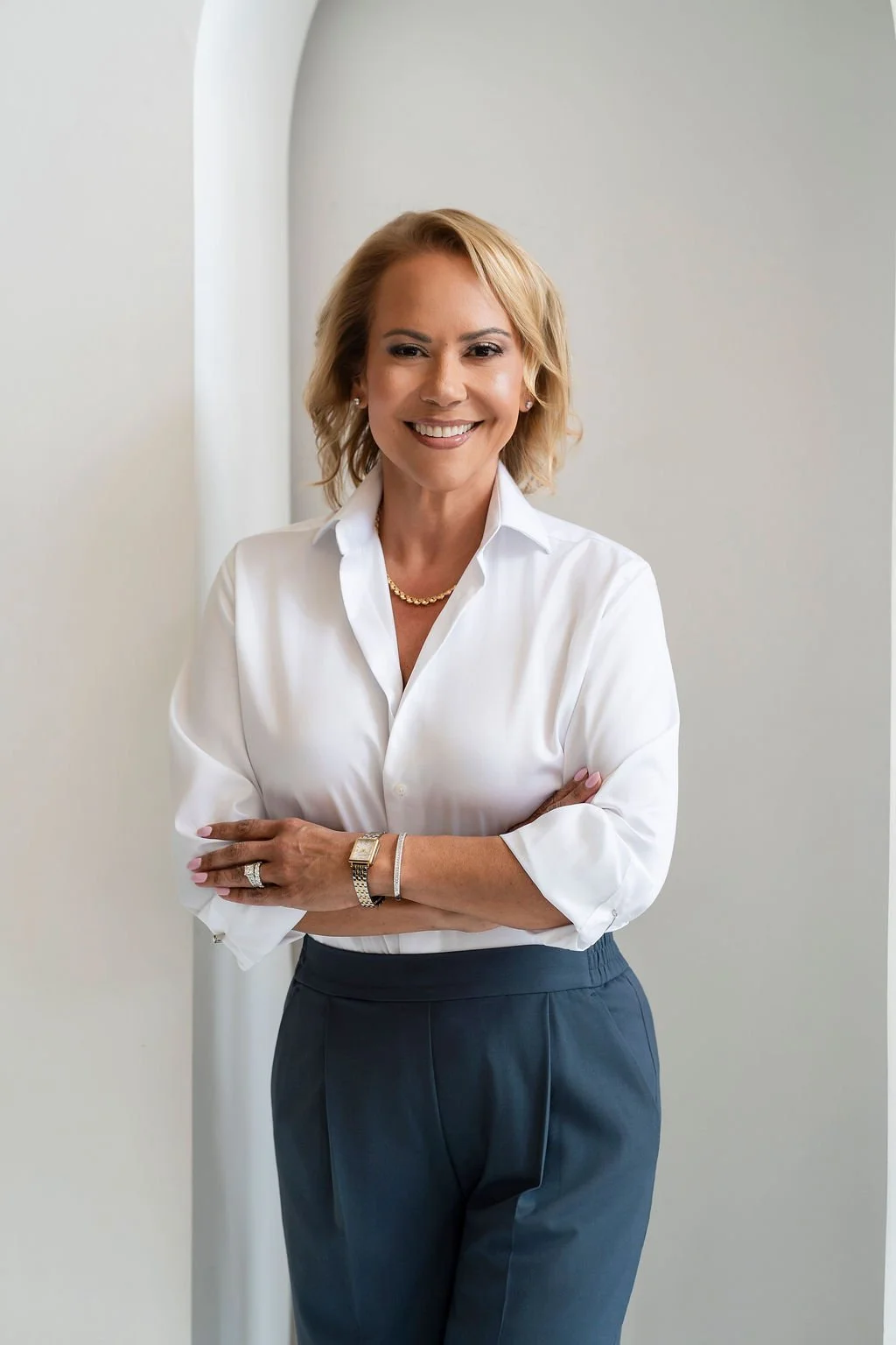 Dr. Dias with short, blonde, curled hair, wearing a white dress shirt and dark blue dress bants, crossing her arms smiling at the camera