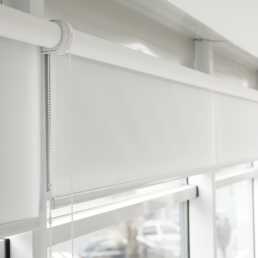 Close-up of a white roller window blind with a pull chain, next to a window with a blurred background of trees and possibly parked cars.
