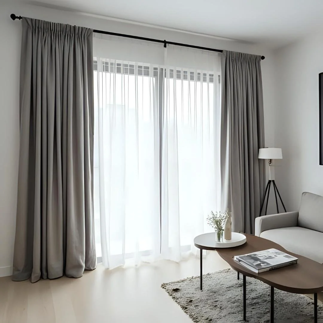 Living room with sheer white curtains and beige drapes on a large window, white sofa, wooden coffee table with magazines and a vase with flowers, floor lamp, and a rug on light-colored floor.