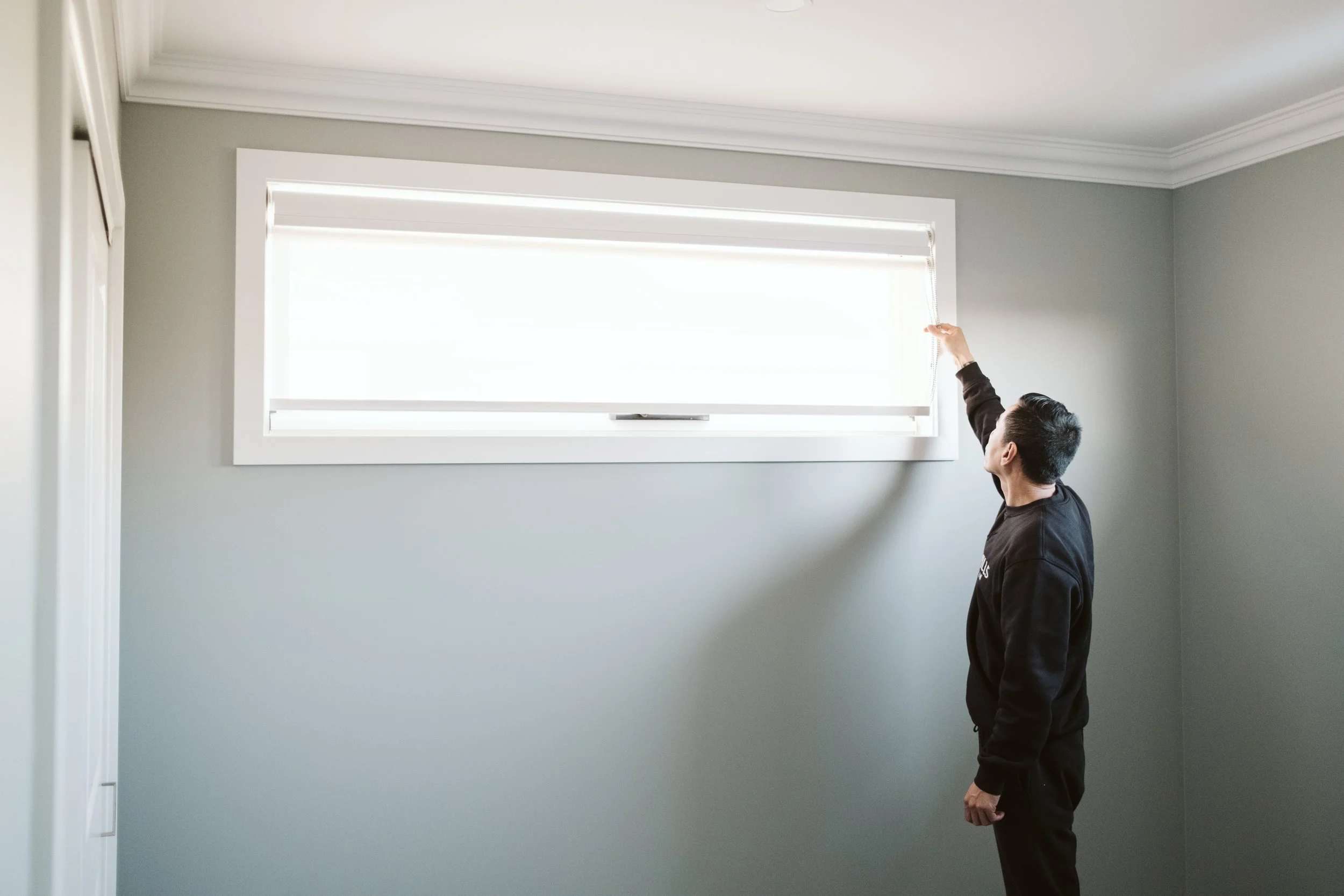 A person adjusting a white horizontal window blind in a room with gray walls and white crown molding.