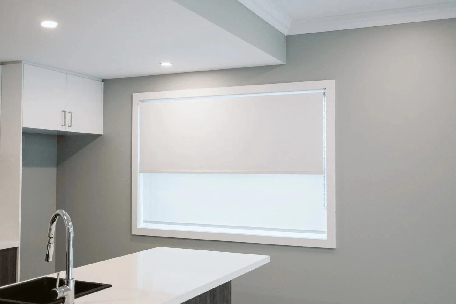 A modern kitchen with white cabinetry, a gray wall, a large window with a white roller blind, and a white countertop with a black sink and silver faucet.