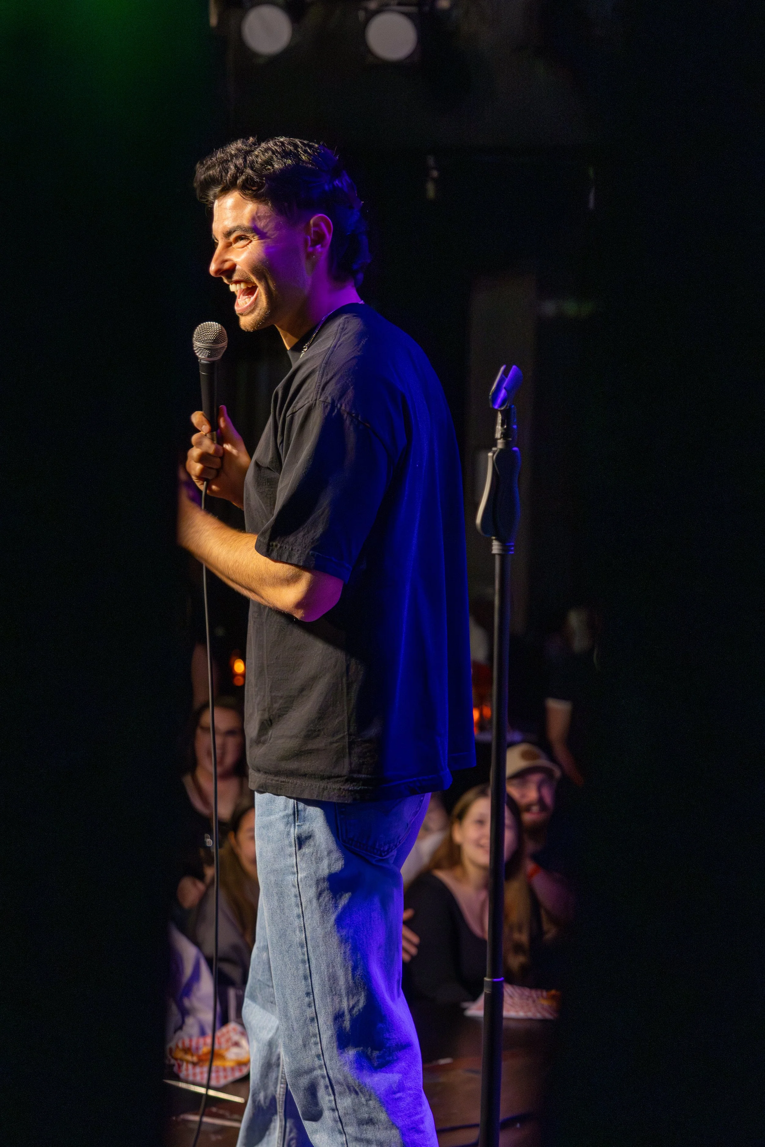 A man with dark hair and a beard, wearing a black shirt and jeans, is on stage holding a microphone and laughing. Audience members are visible in the background, watching him perform.