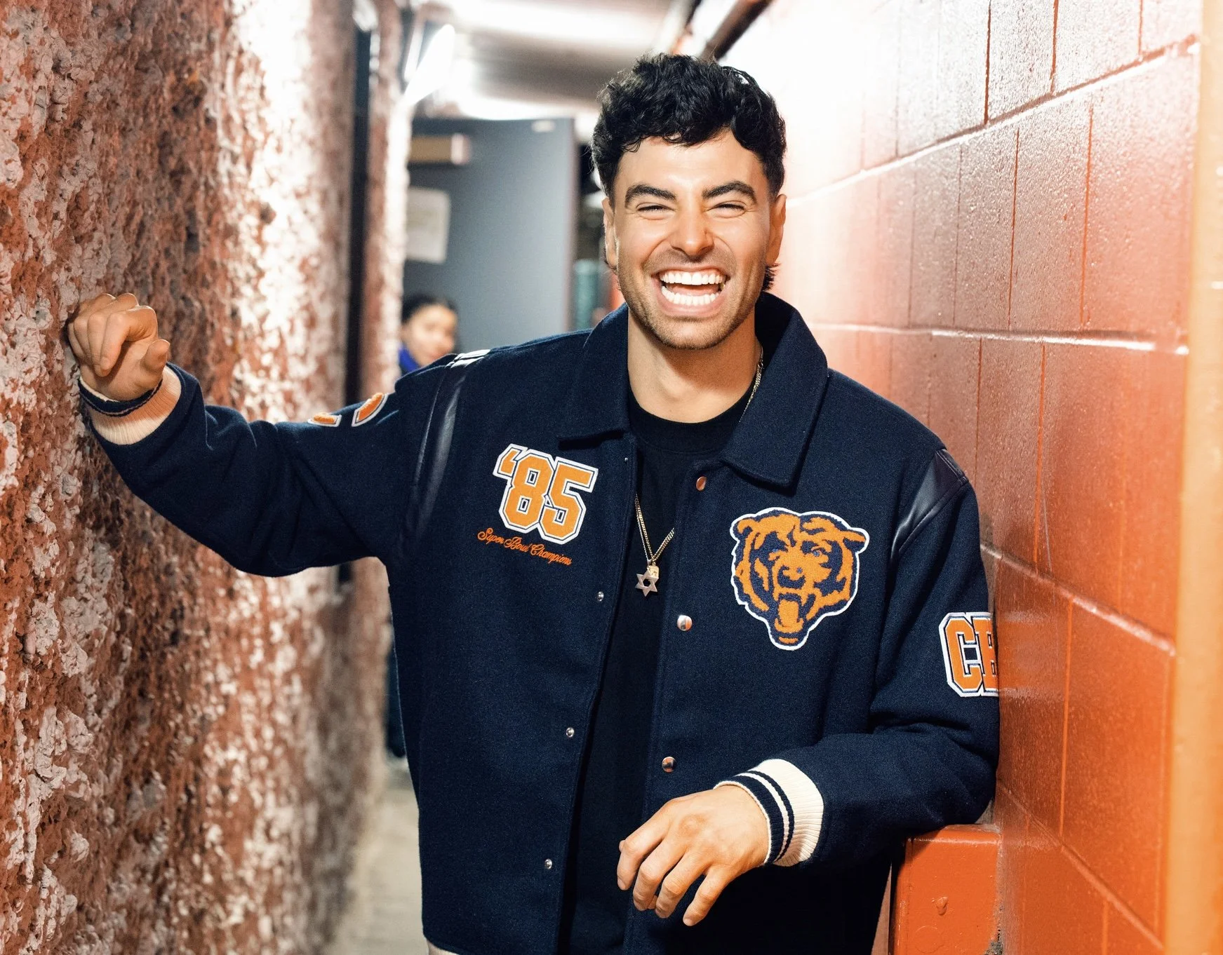 A young man with dark, curly hair and a big smile, wearing a navy blue varsity jacket with orange and white patches, standing in a narrow corridor with textured brick walls, making a fist gesture with his right hand.