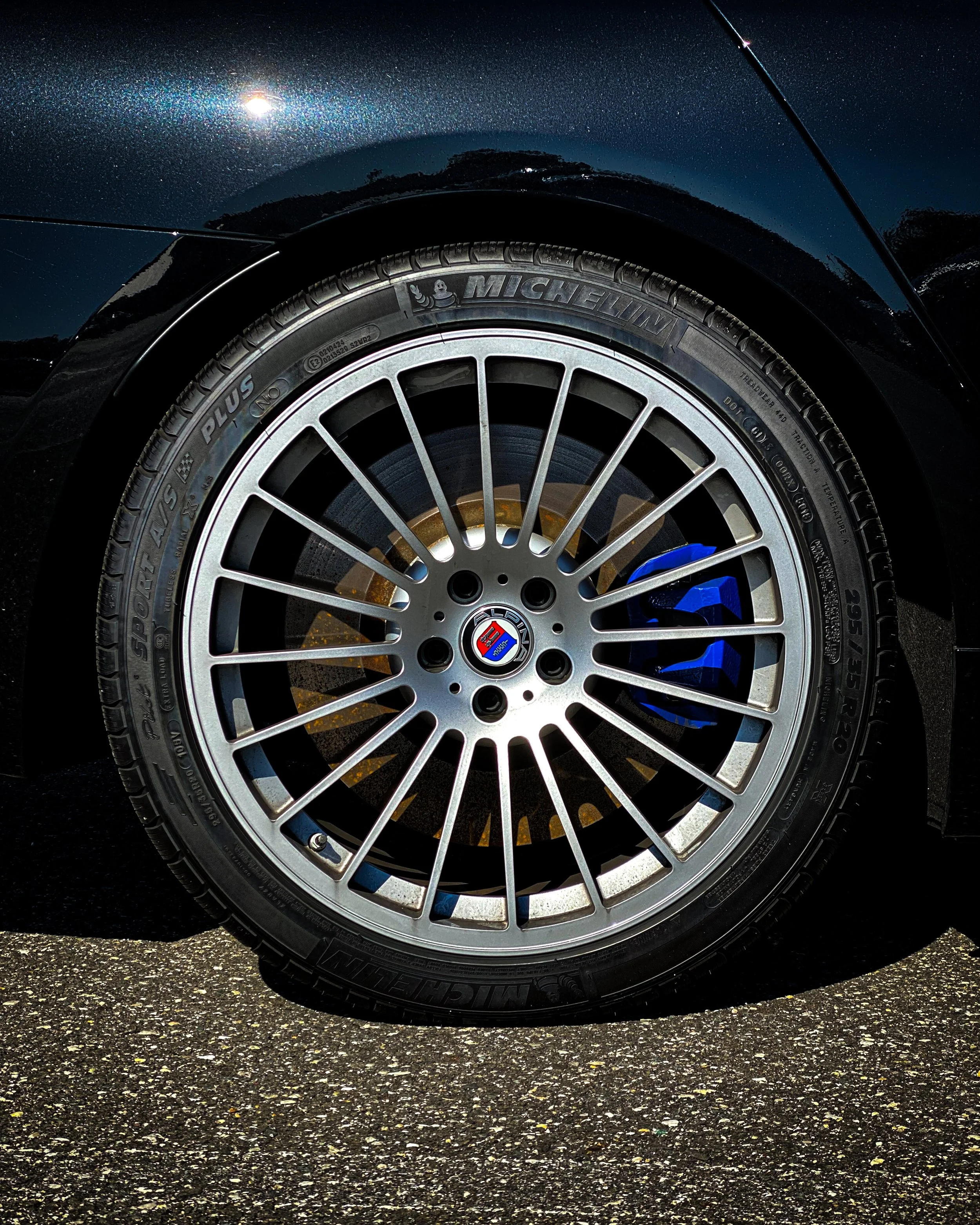 Close-up of a car tire and wheel on asphalt, with a dark blue body and a reflection of the sky and trees.