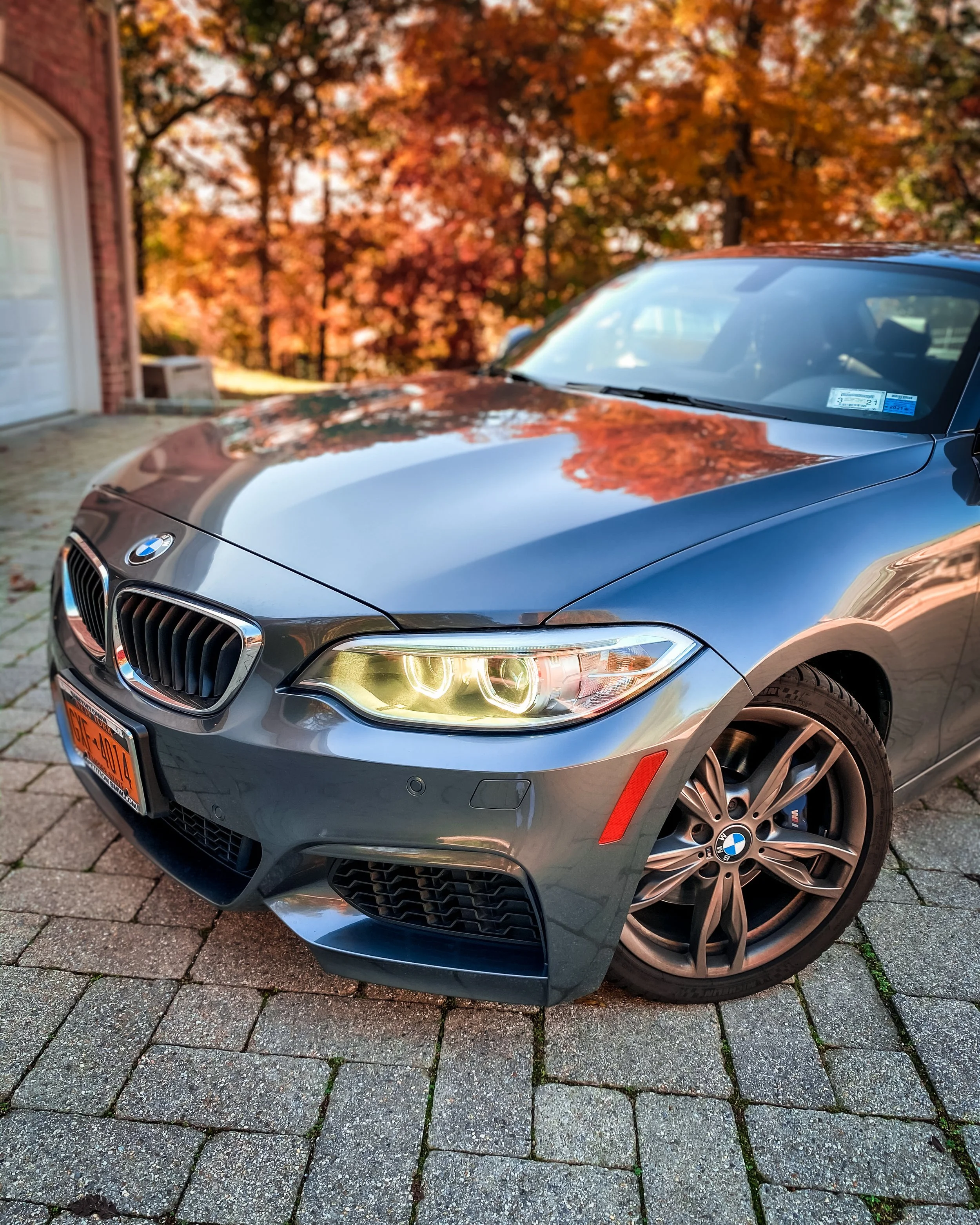 A close-up view of a gray BMW car parked on a stone driveway with autumn orange and red trees in the background.