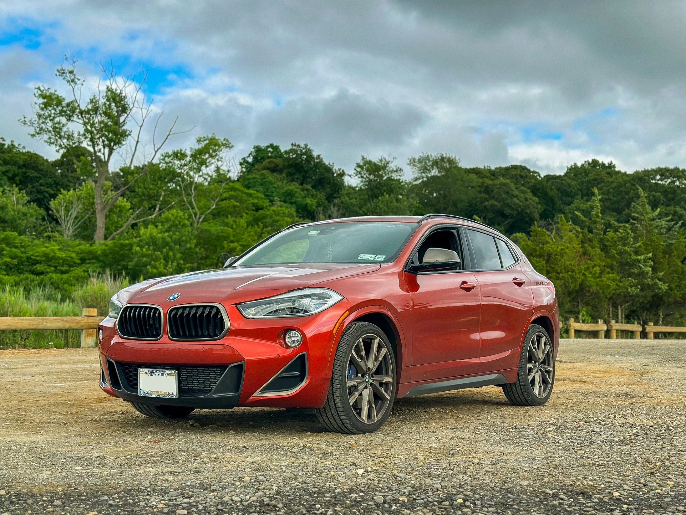 Red BMW SUV parked on a gravel lot with lush green trees and a cloudy sky in the background.