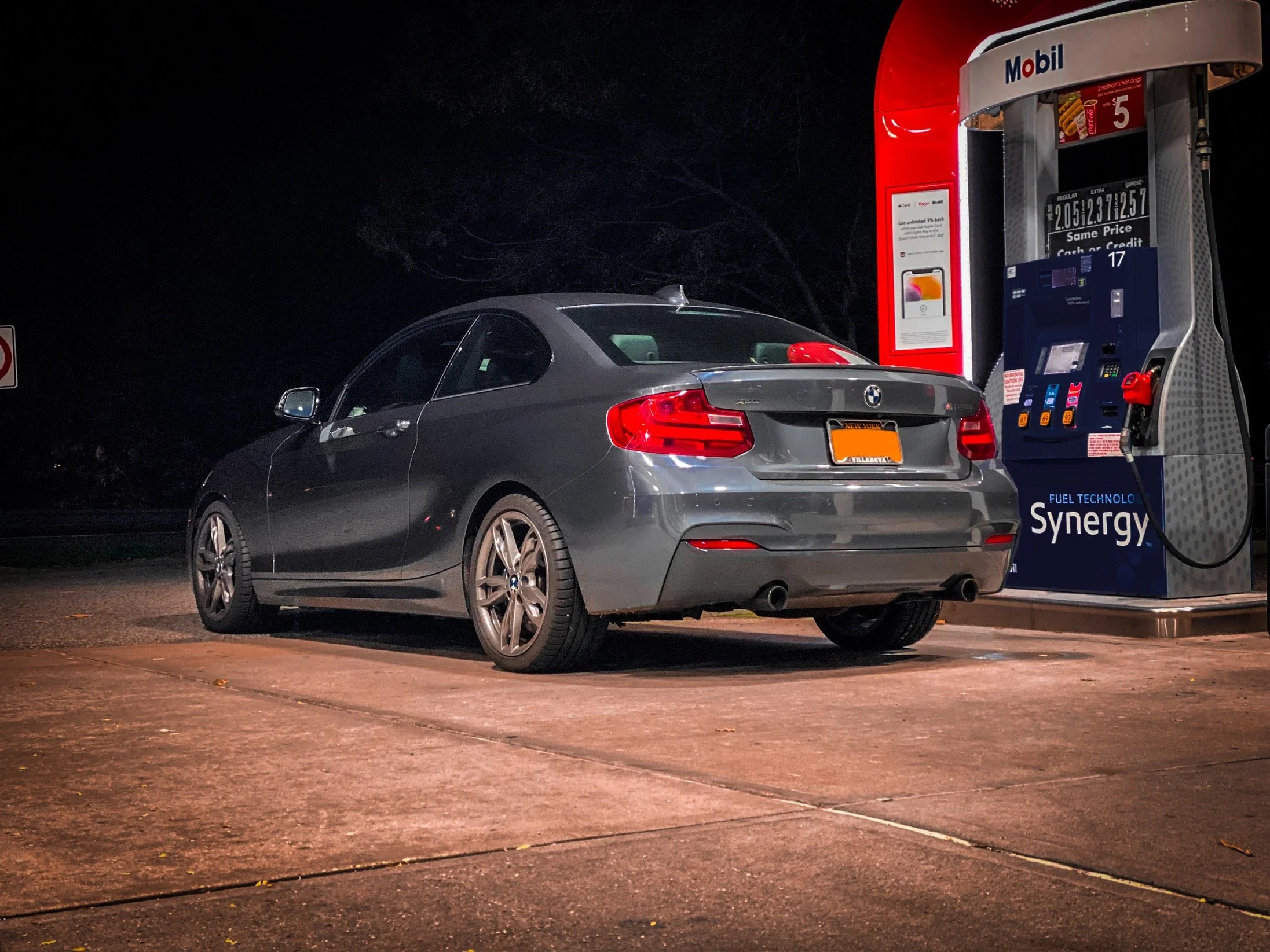 A gray BMW car at a Mobil gas station at night with an illuminated pump and black sky in the background.