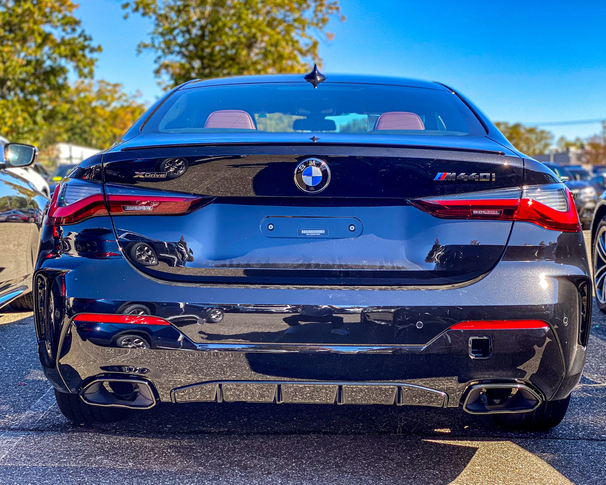 The rear view of a black BMW M460i car with BMW and M logos, parked in a lot with other vehicles, under a partly cloudy sky with trees in the background.