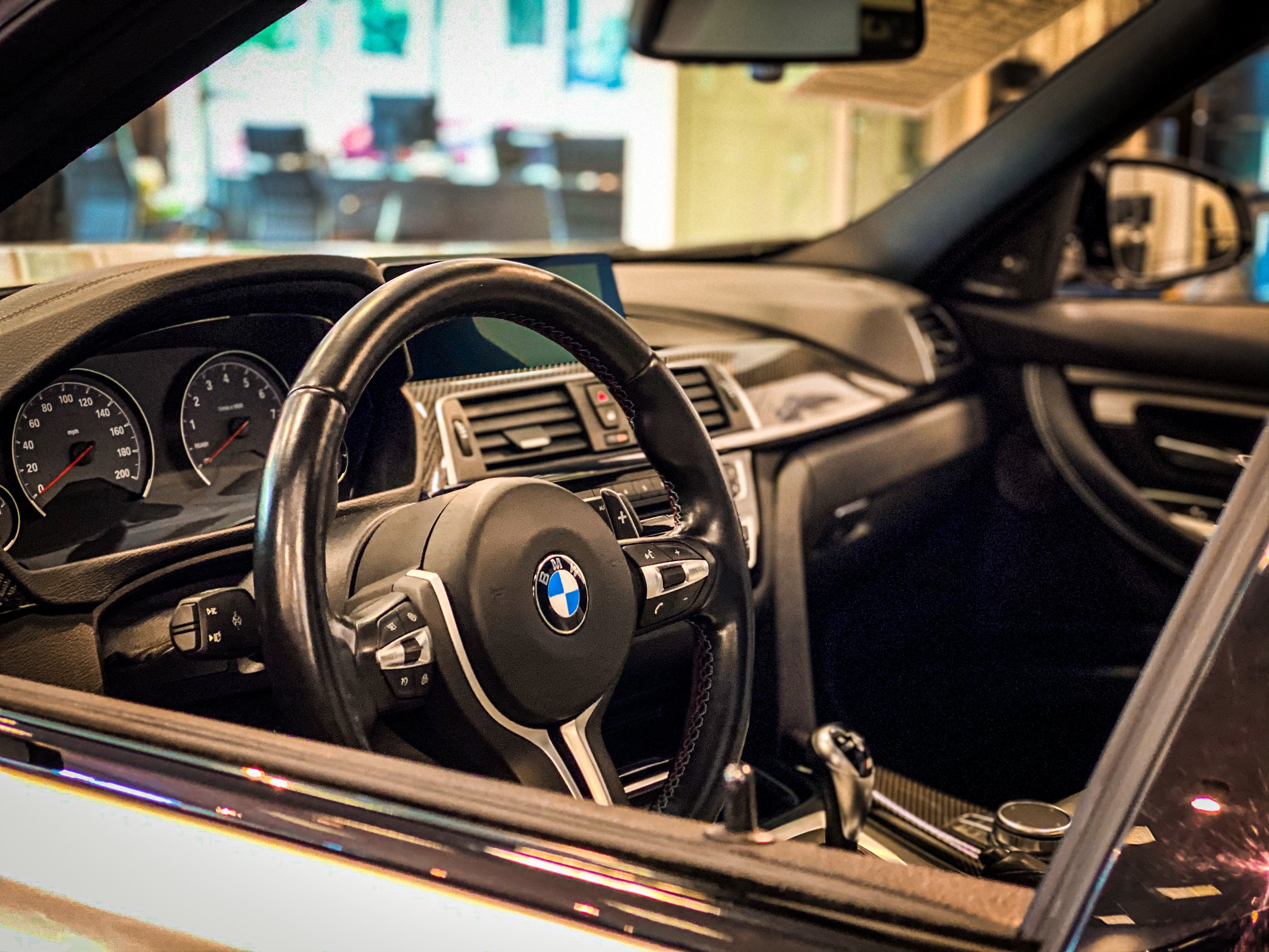 Interior view of a BMW vehicle showing the steering wheel, dashboard, and instrument gauges, with a blurred background of what appears to be a garage or workshop.