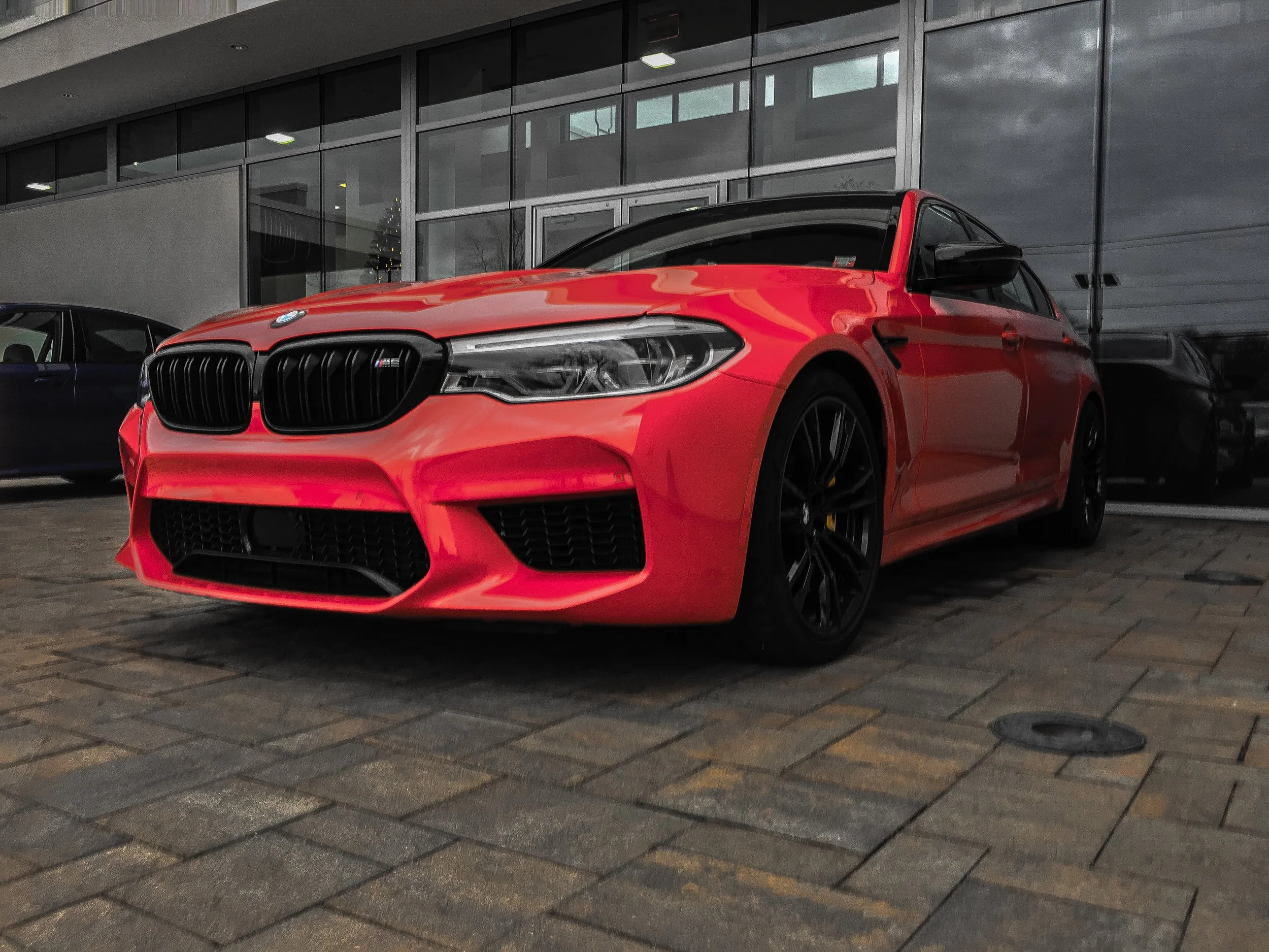 Red BMW M8 parked on a city street near a building with large glass windows at night.