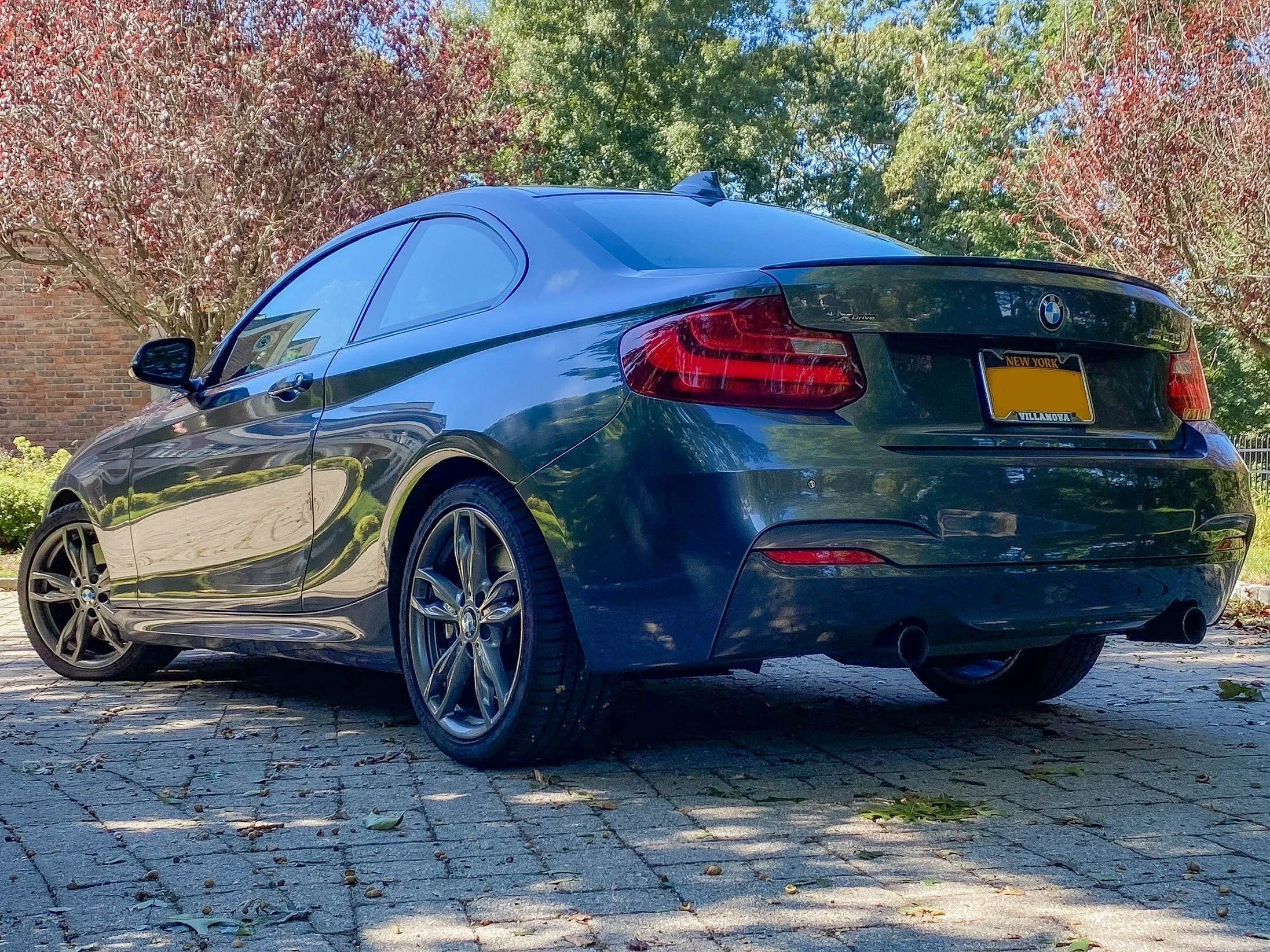 A blue BMW convertible car parked on a cobblestone driveway surrounded by trees with green and reddish leaves.