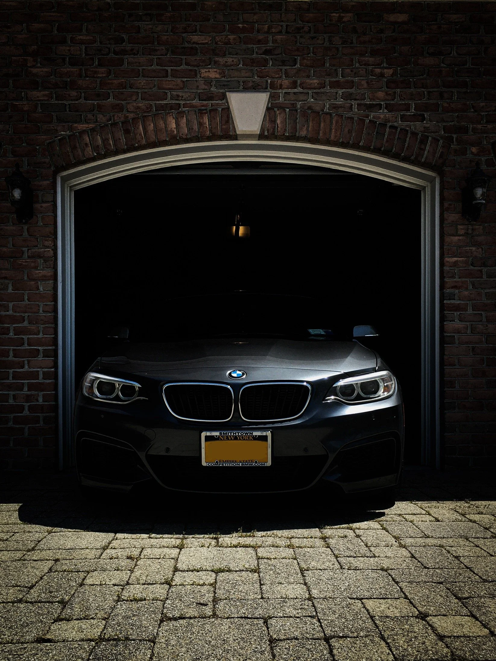 A silver BMW car parked inside a garage with a brick exterior and a black interior, showing front headlights, grille, and license plate.