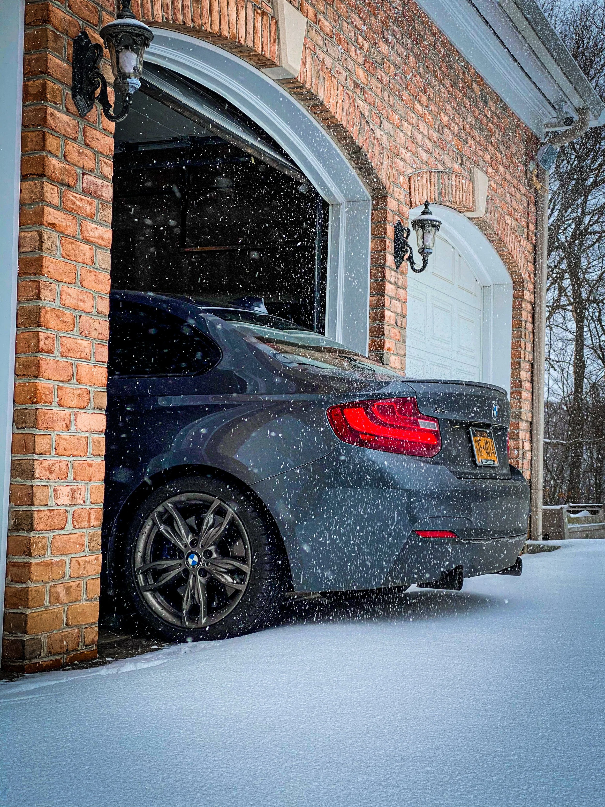 A gray BMW parked in a garage with brick walls and exterior-mounted vintage-style lanterns, snow falling and covering the ground.