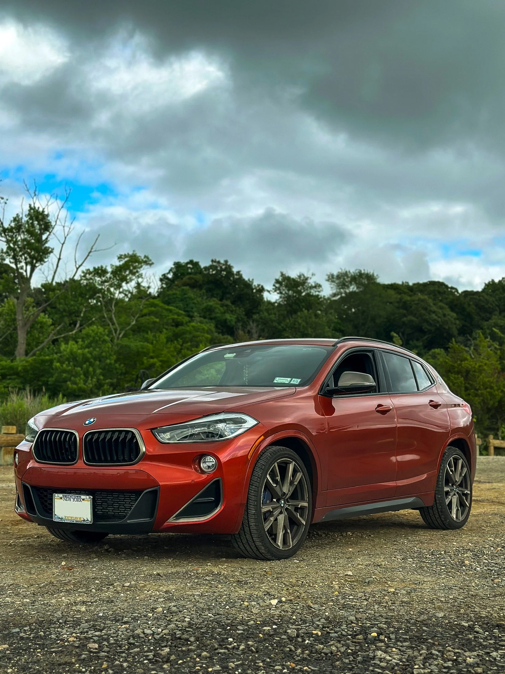 A red BMW SUV parked on a gravel surface with green trees and cloudy sky in the background.