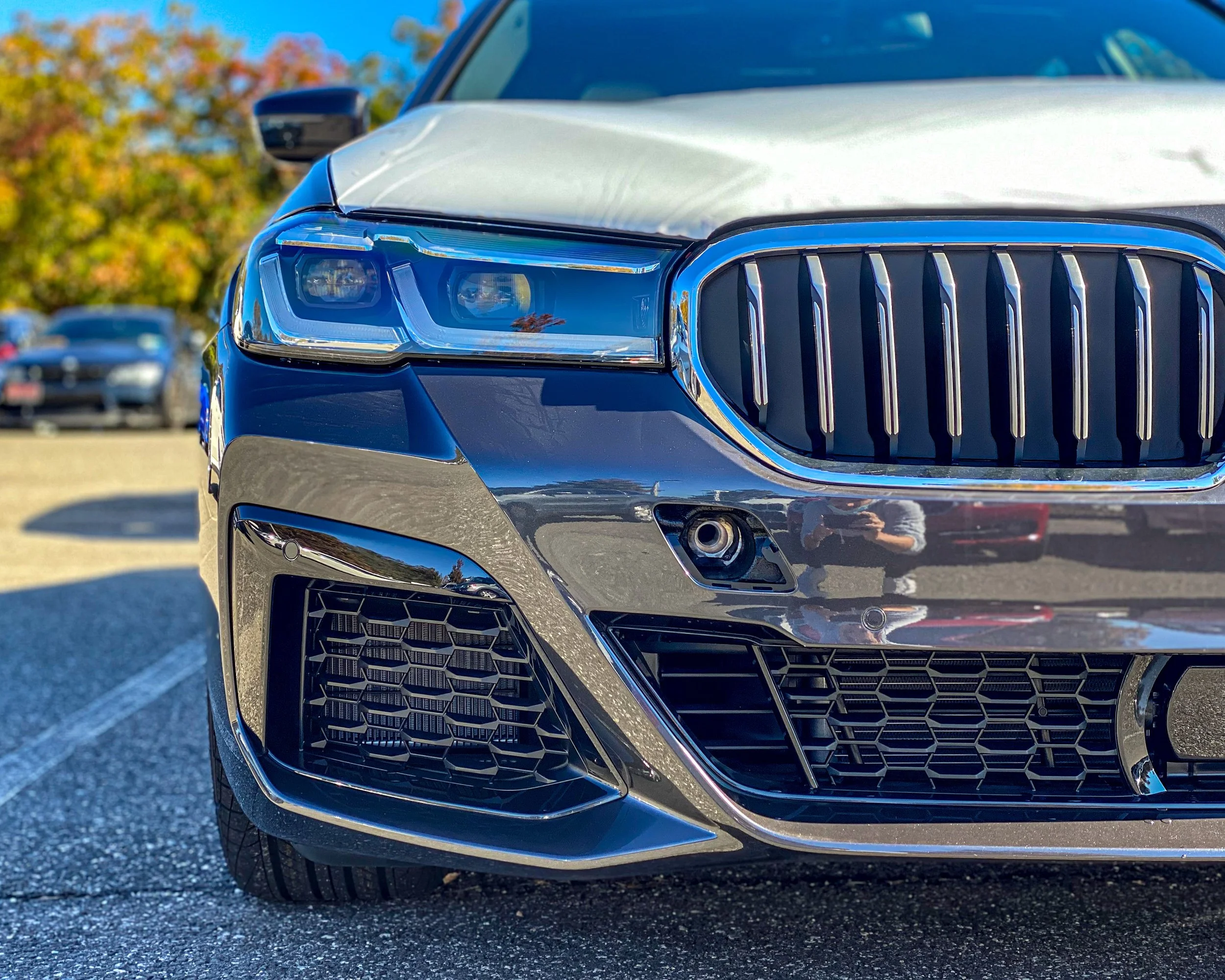 Close-up of the front of a gray luxury car, showing the headlight, grille, and bumper in an outdoor parking lot.
