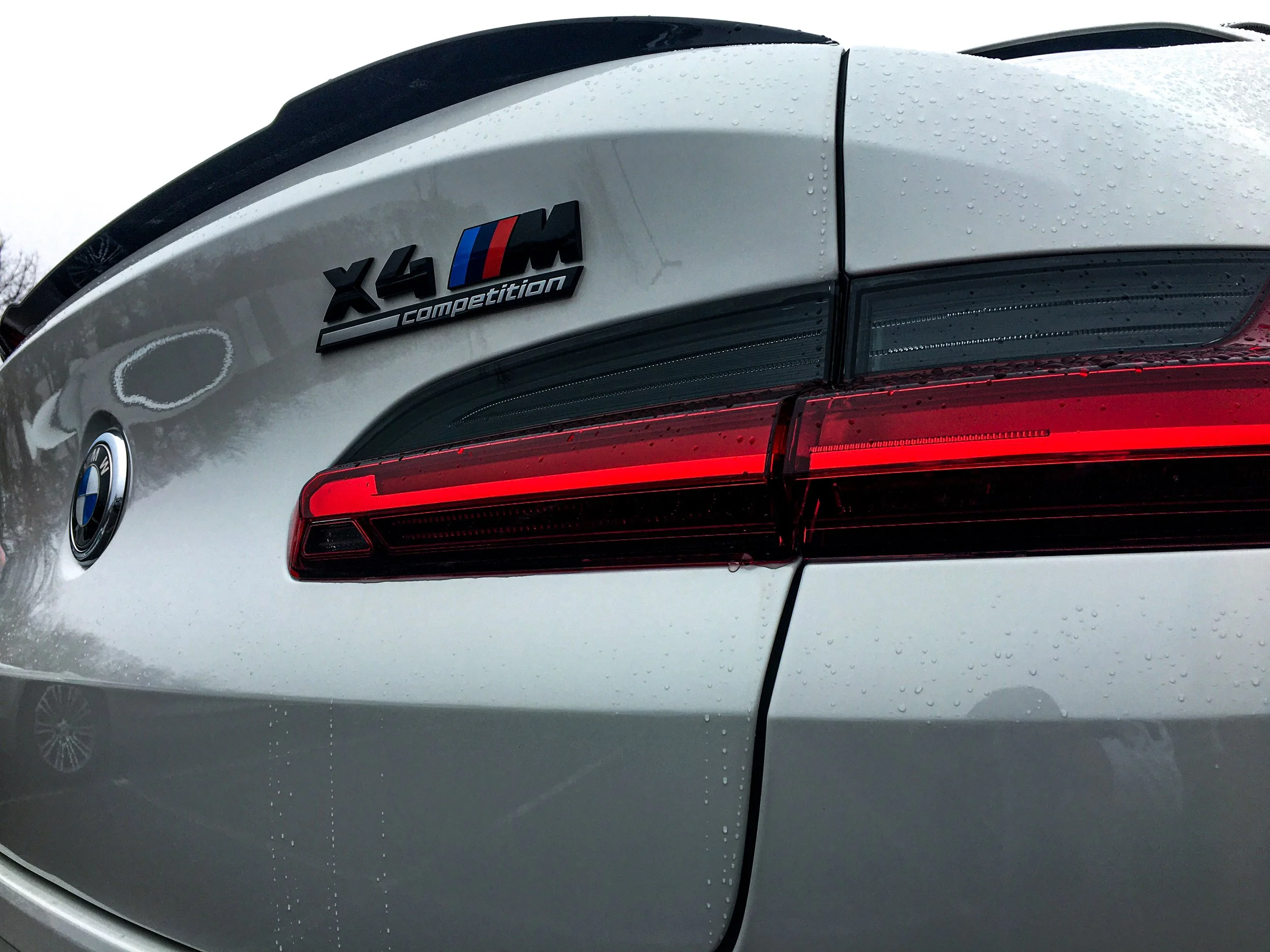 Close-up view of the rear part of a white BMW X4 M Competition vehicle, showing the BMW badge and M series badge, with some raindrops on the surface.
