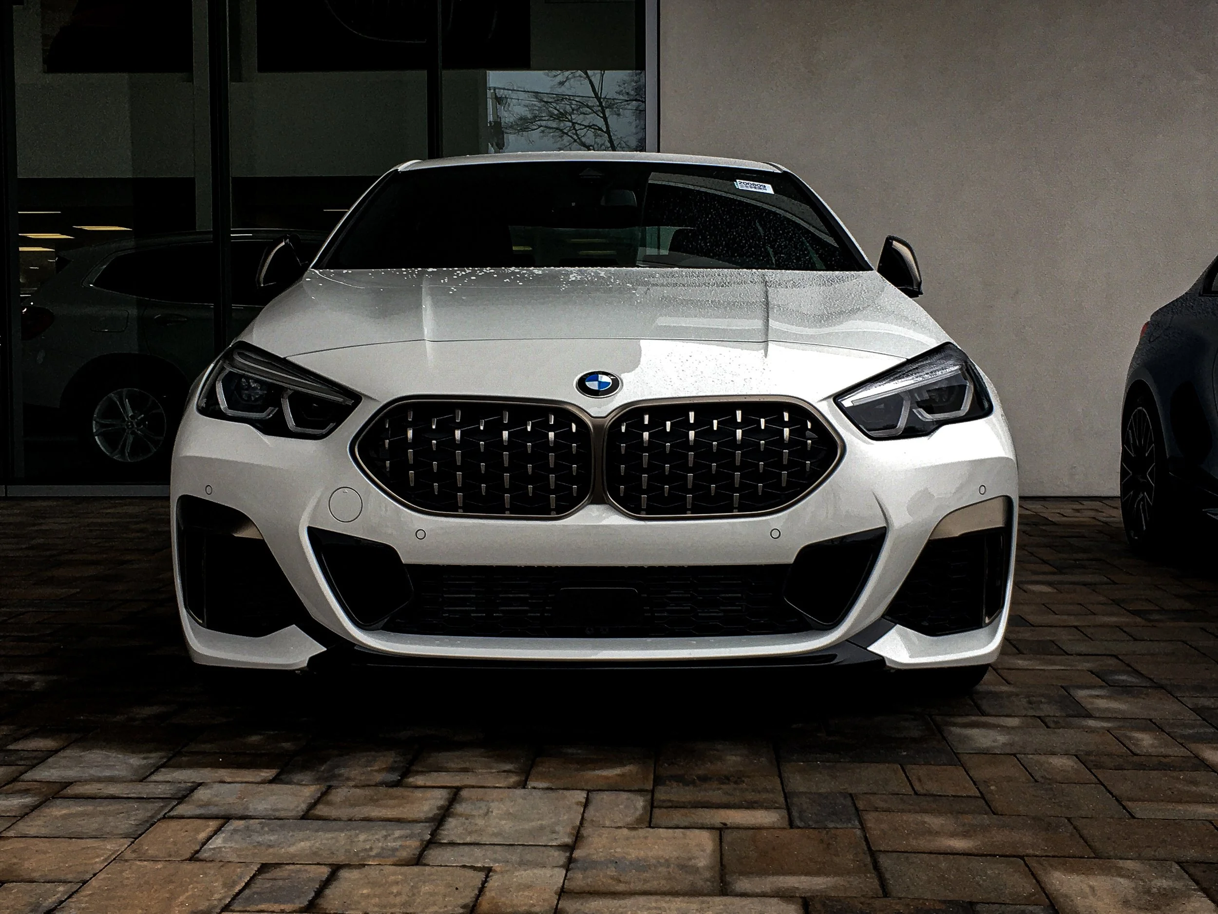 White BMW car parked outside on a brick pavement, with a building and reflective windows behind it.