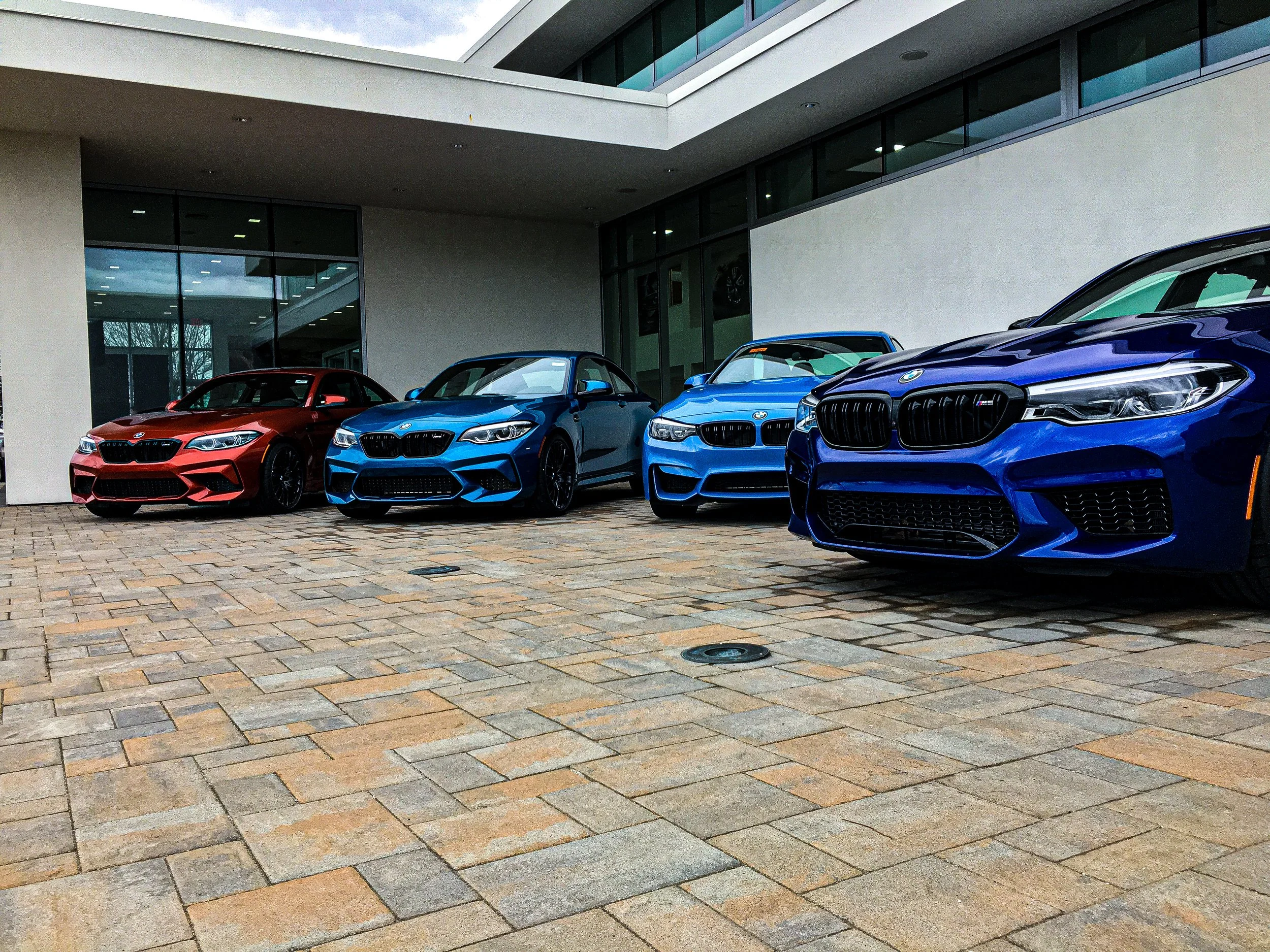 A row of four blue and red BMW cars parked in front of a modern building with large glass windows.