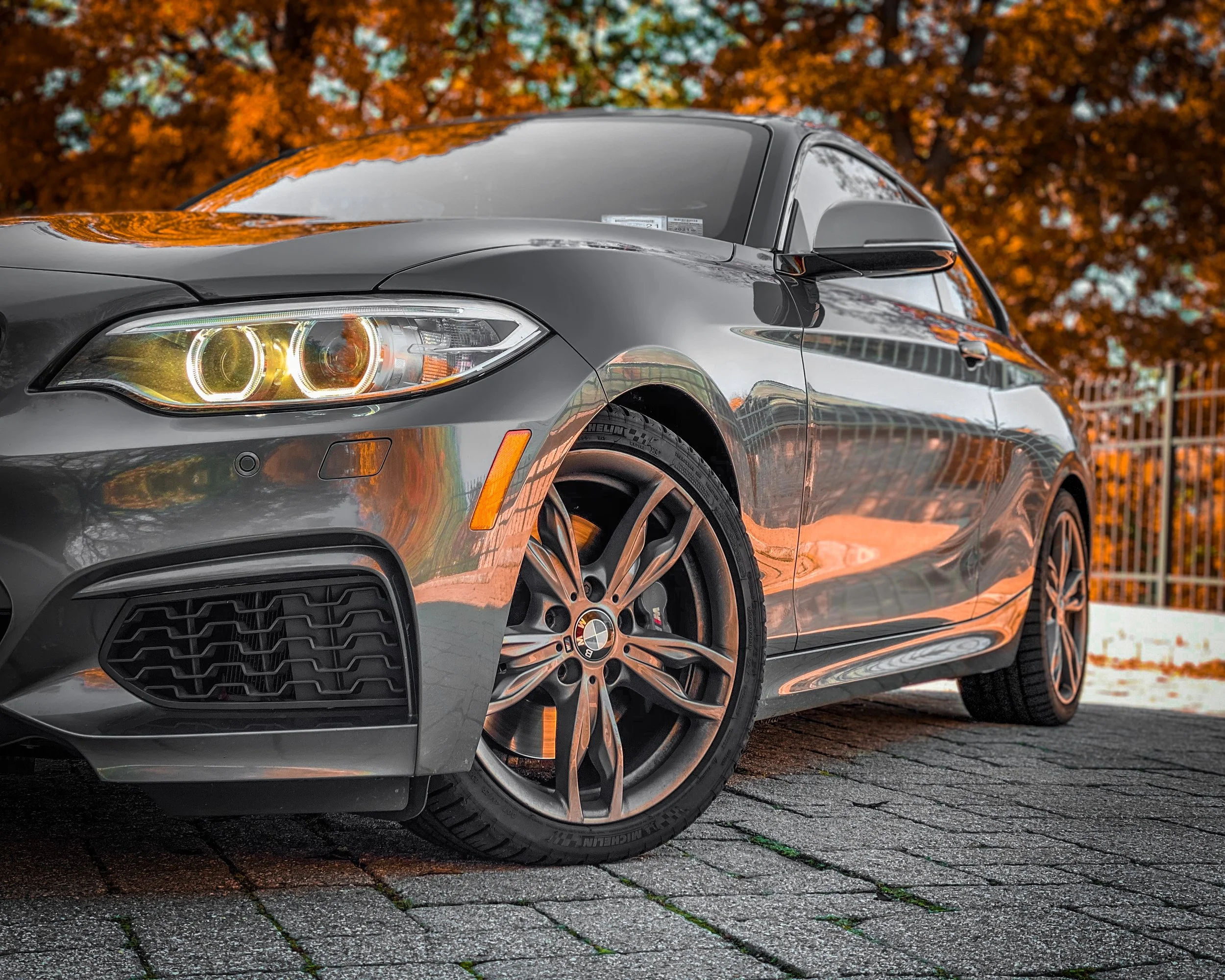A black BMW car parked on a cobblestone street with autumn-colored trees in the background.