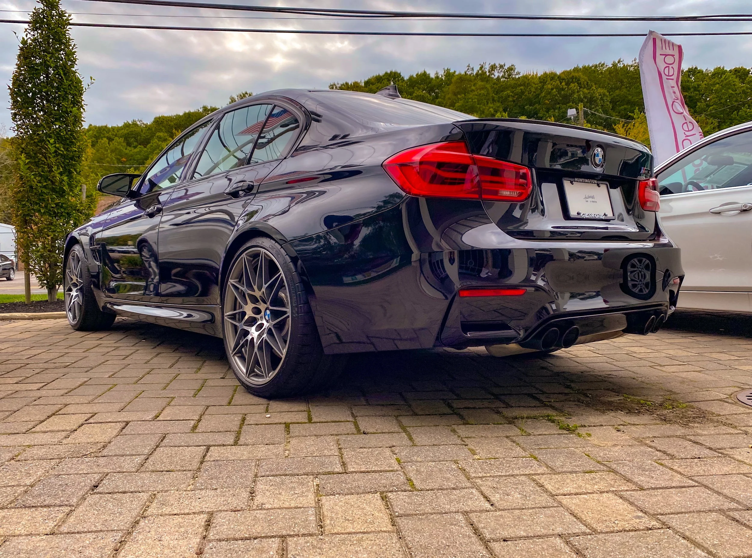 A black BMW sedan parked on a paved area outside, with a white car partially visible beside it, a tree, and green hills in the background under a cloudy sky.