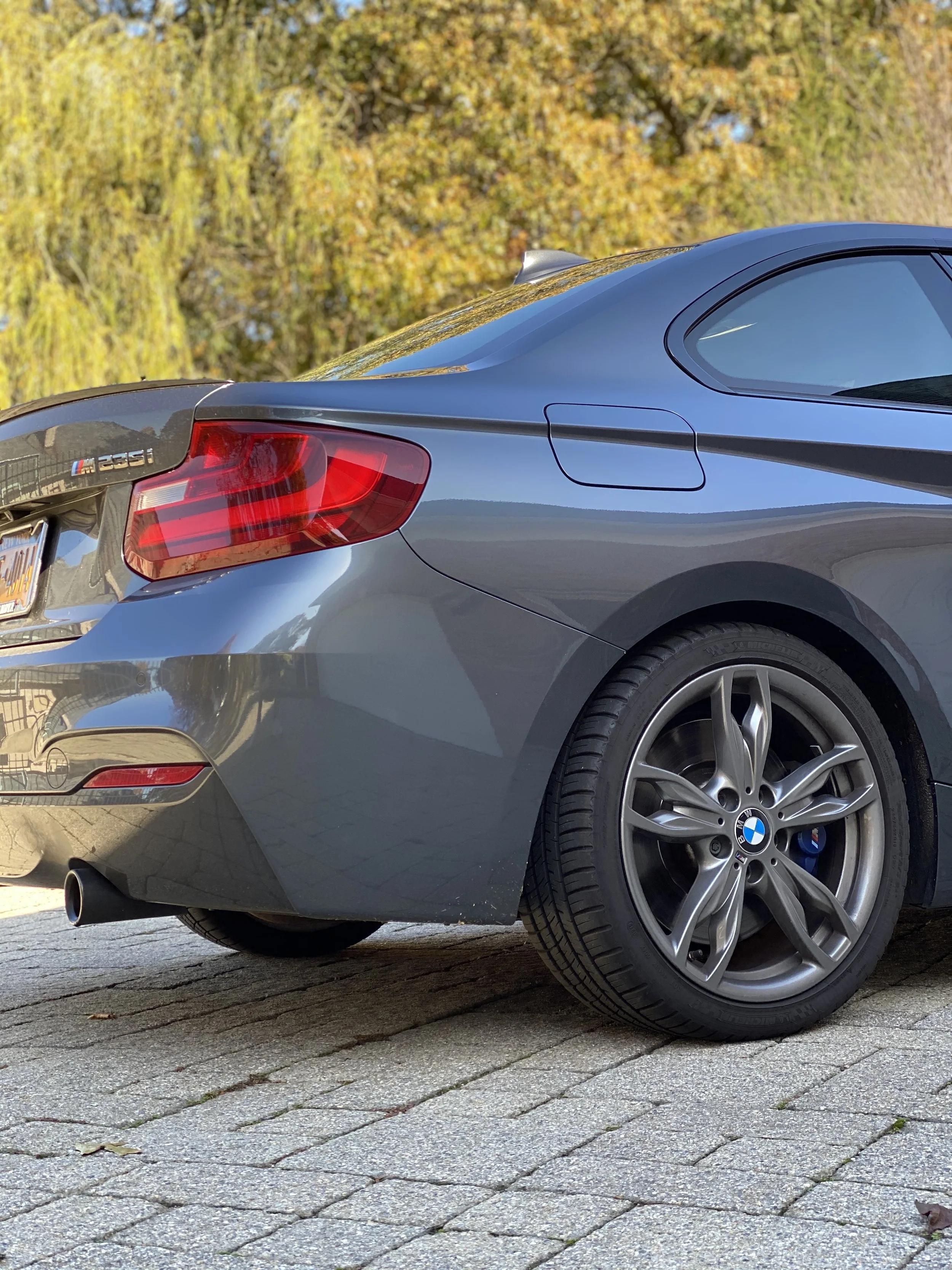 The rear side of a gray BMW M235i car parked on a brick pavement with trees displaying fall foliage in the background.