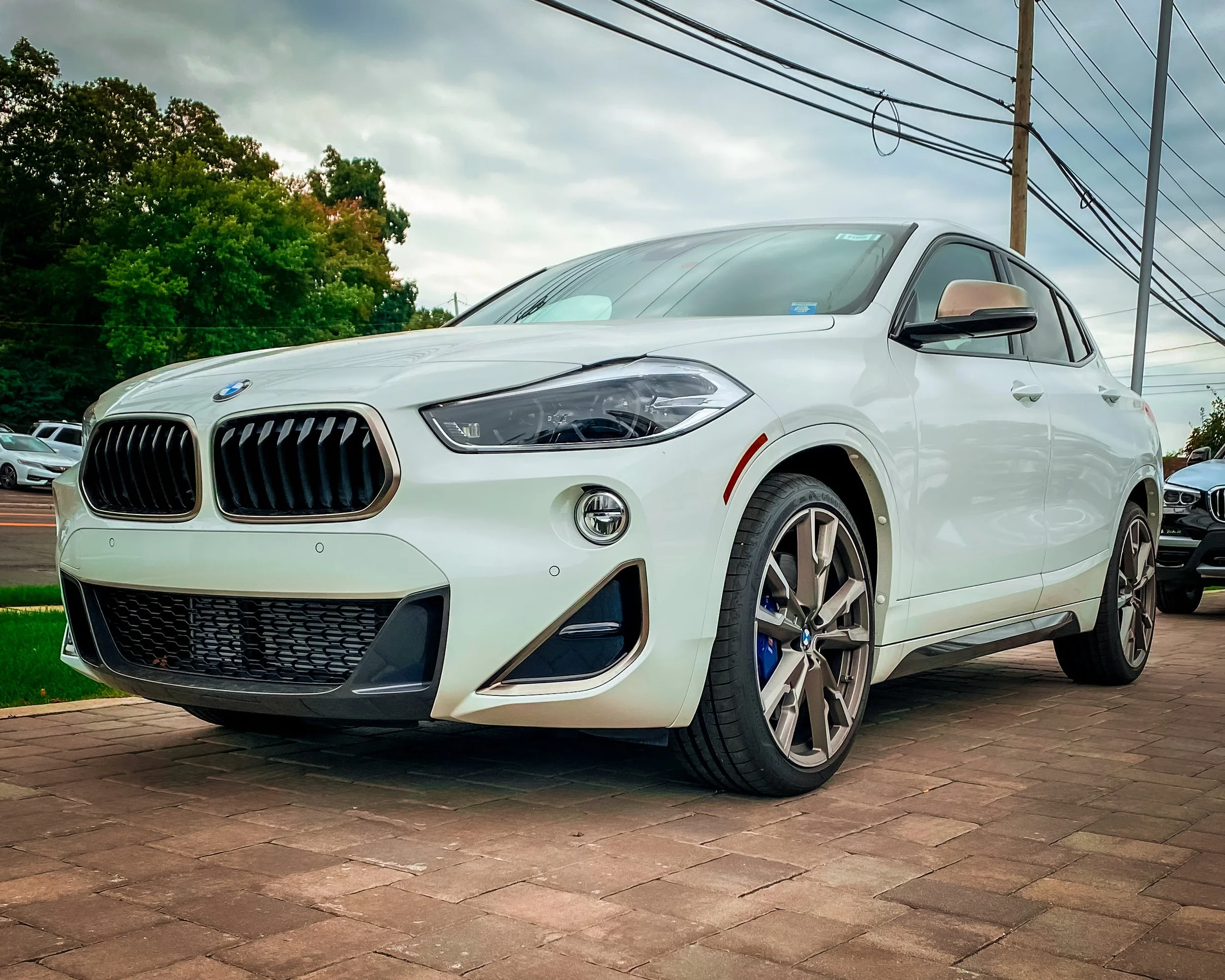 A white BMW SUV parked on a brick pavement with other cars in the background. There are green trees and utility poles with power lines under a cloudy sky.